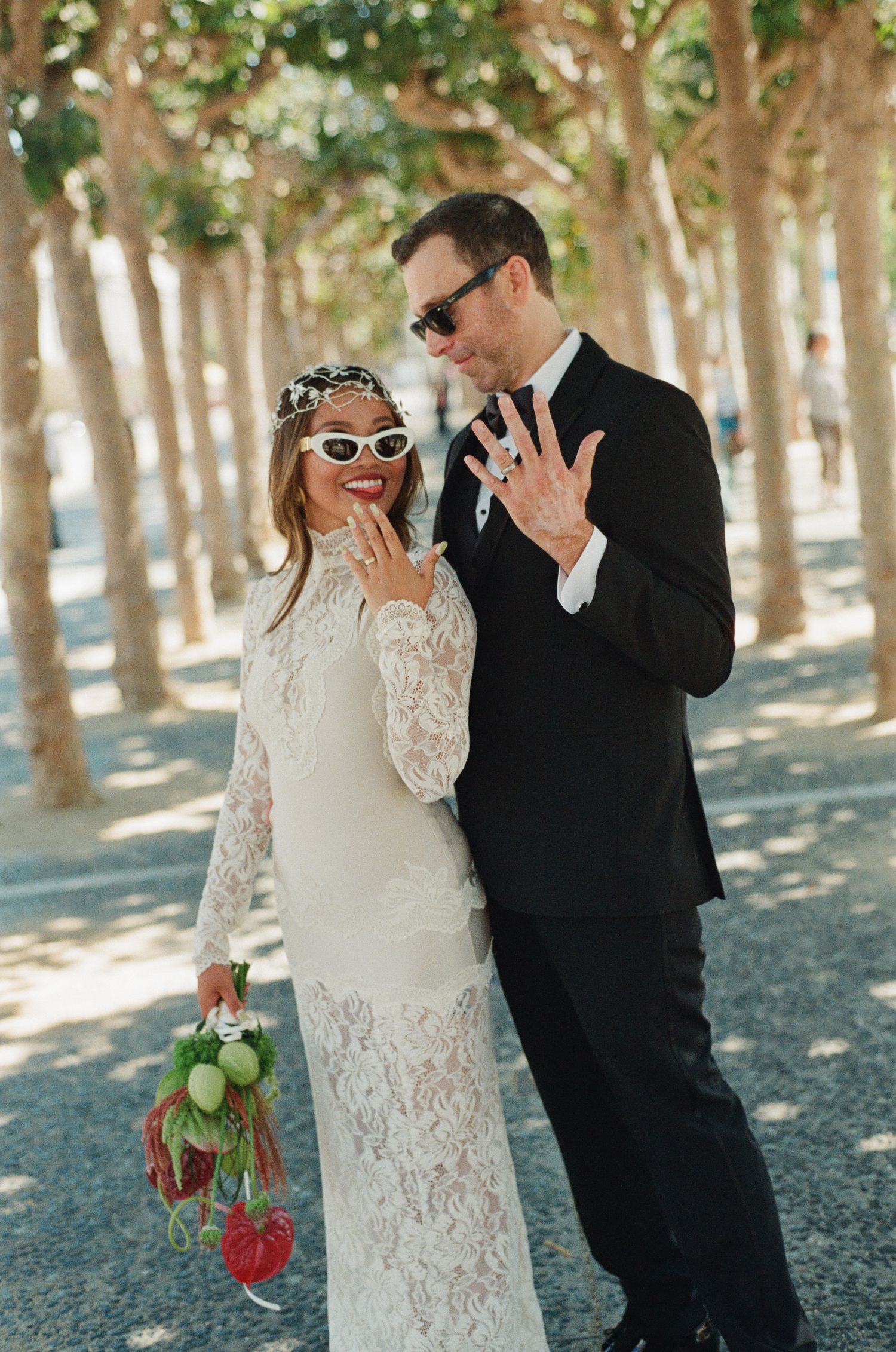 Stylish bride and groom wearing sunglasses pose playfully in a tree-lined path on their wedding day.