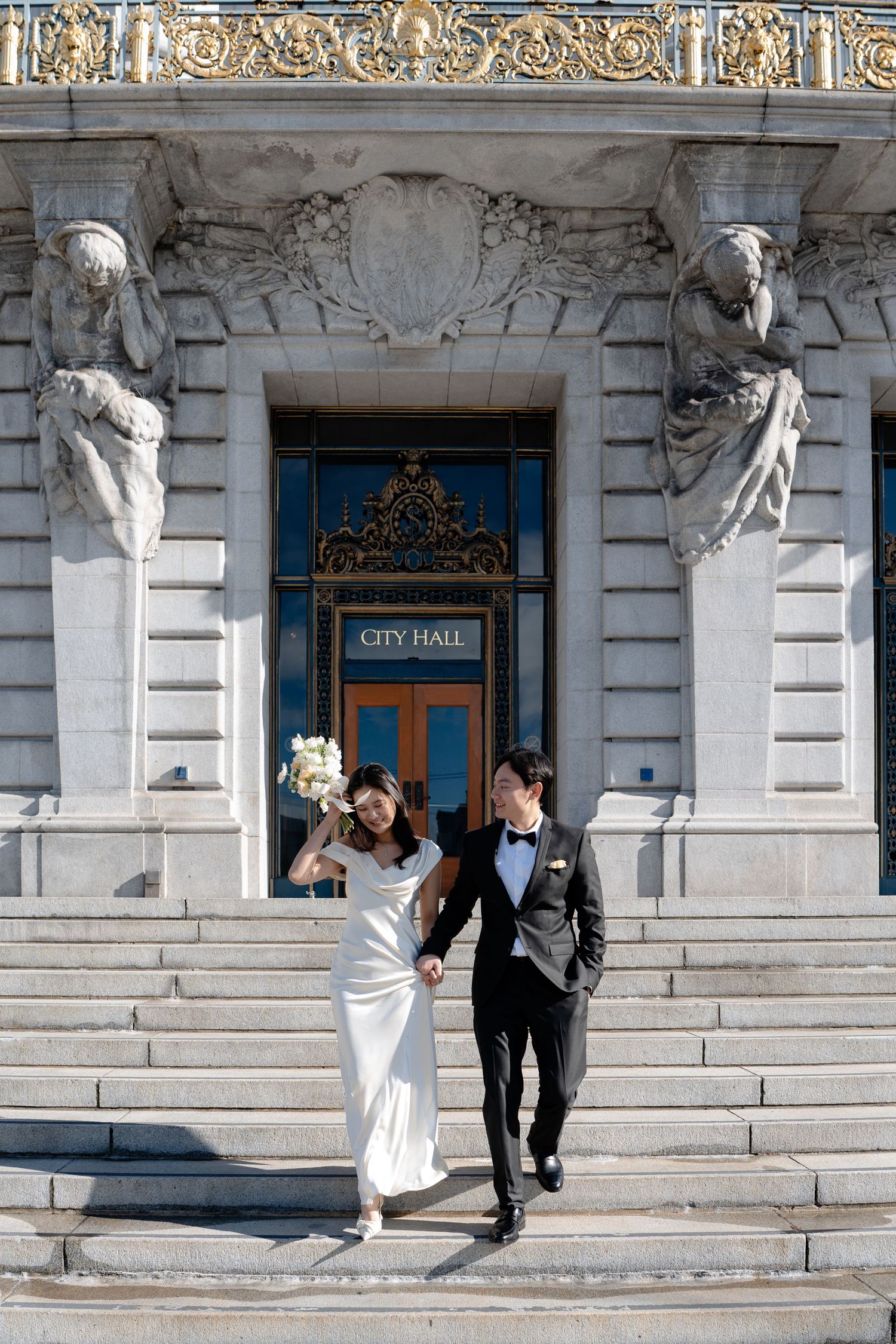Newlywed couple poses playfully on grand marble steps of historic City Hall building during wedding photoshoot.