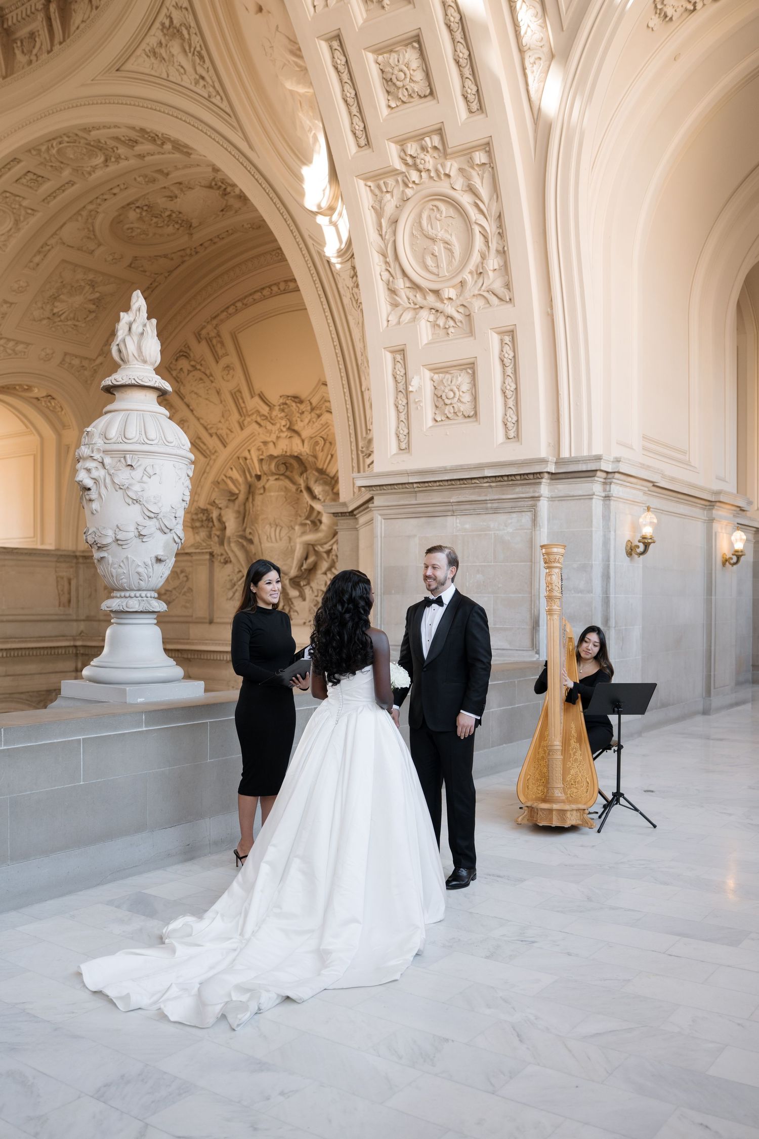 Small wedding ceremony takes place in an elegant marble hallway with ornate architectural details and decorative urn.
