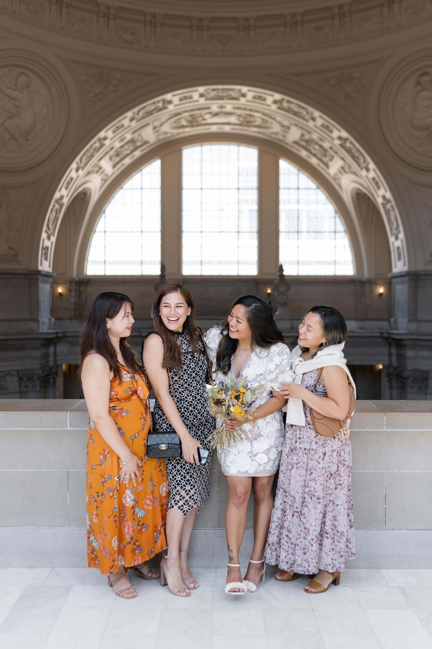 A group of friends wearing floral dresses laugh together under an ornate arched ceiling at San Francisco City Hall.