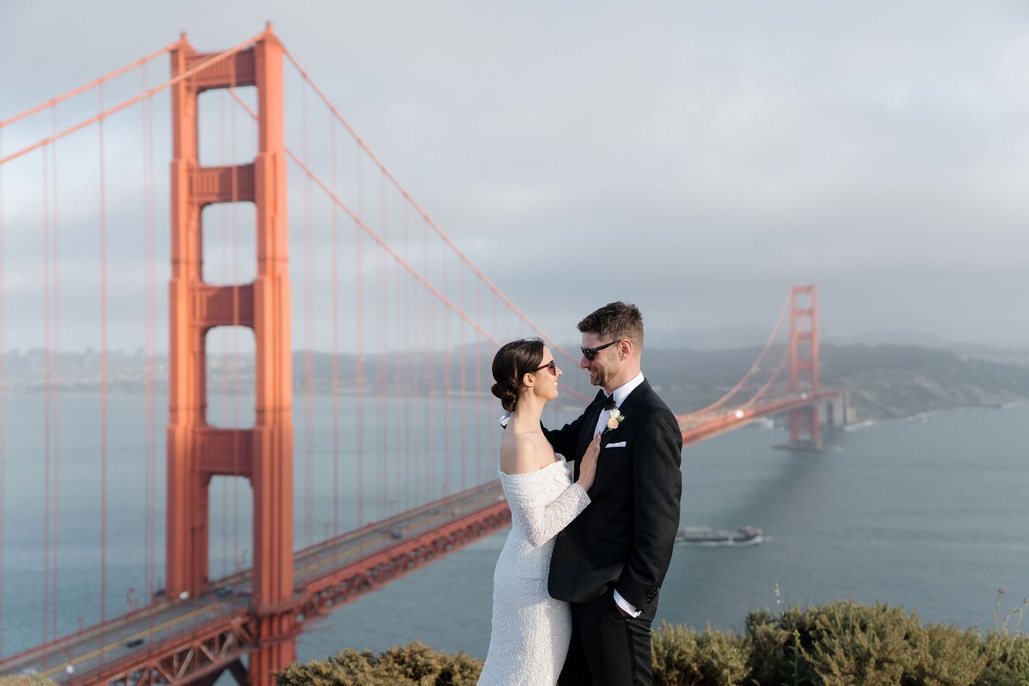 A newlywed couple stands on an overlook by the iconic Golden Gate Bridge in San Francisco on a cloudy day.