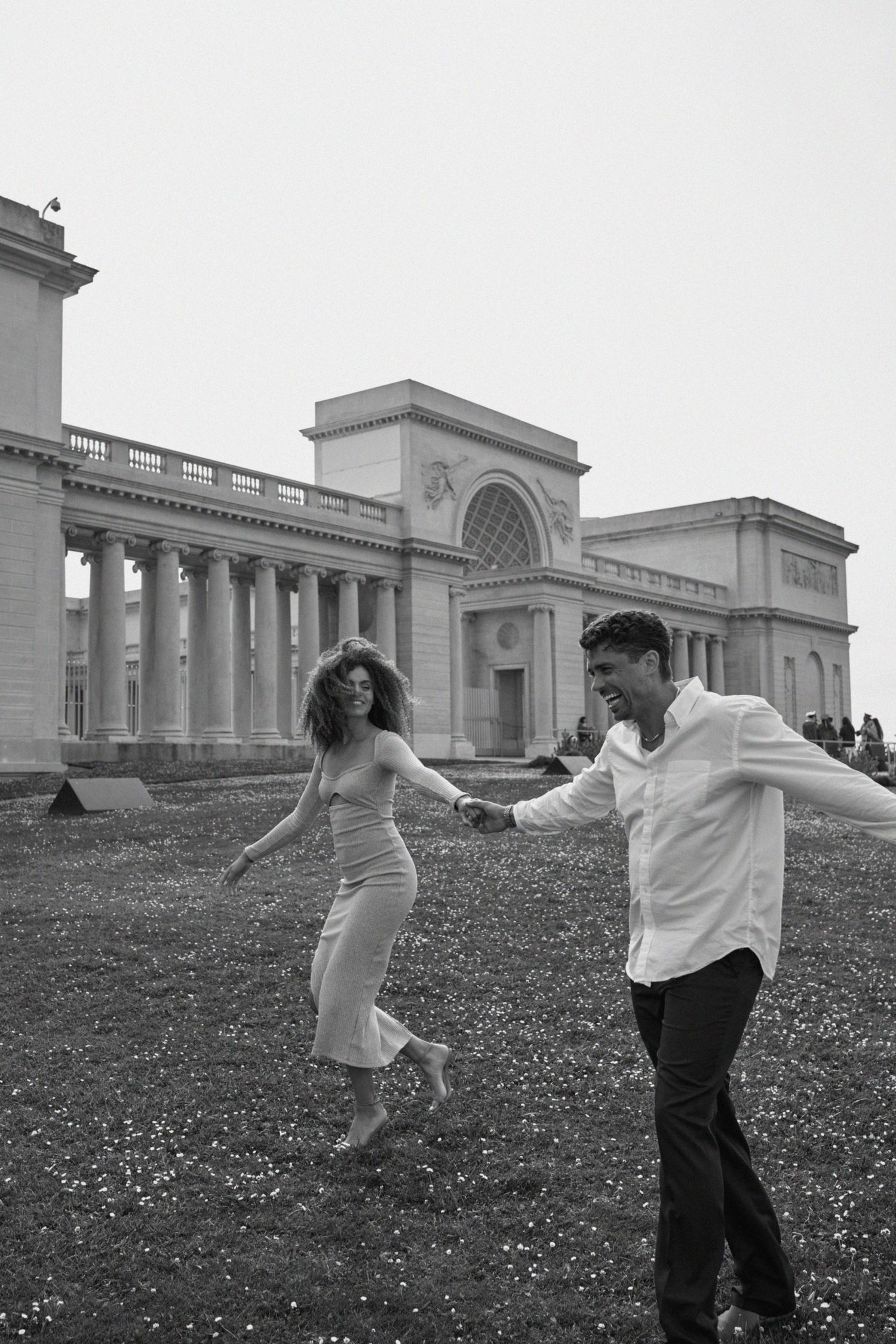 A joyful couple dances together on a grassy lawn in front of a grand neoclassical building with columns and arches.
