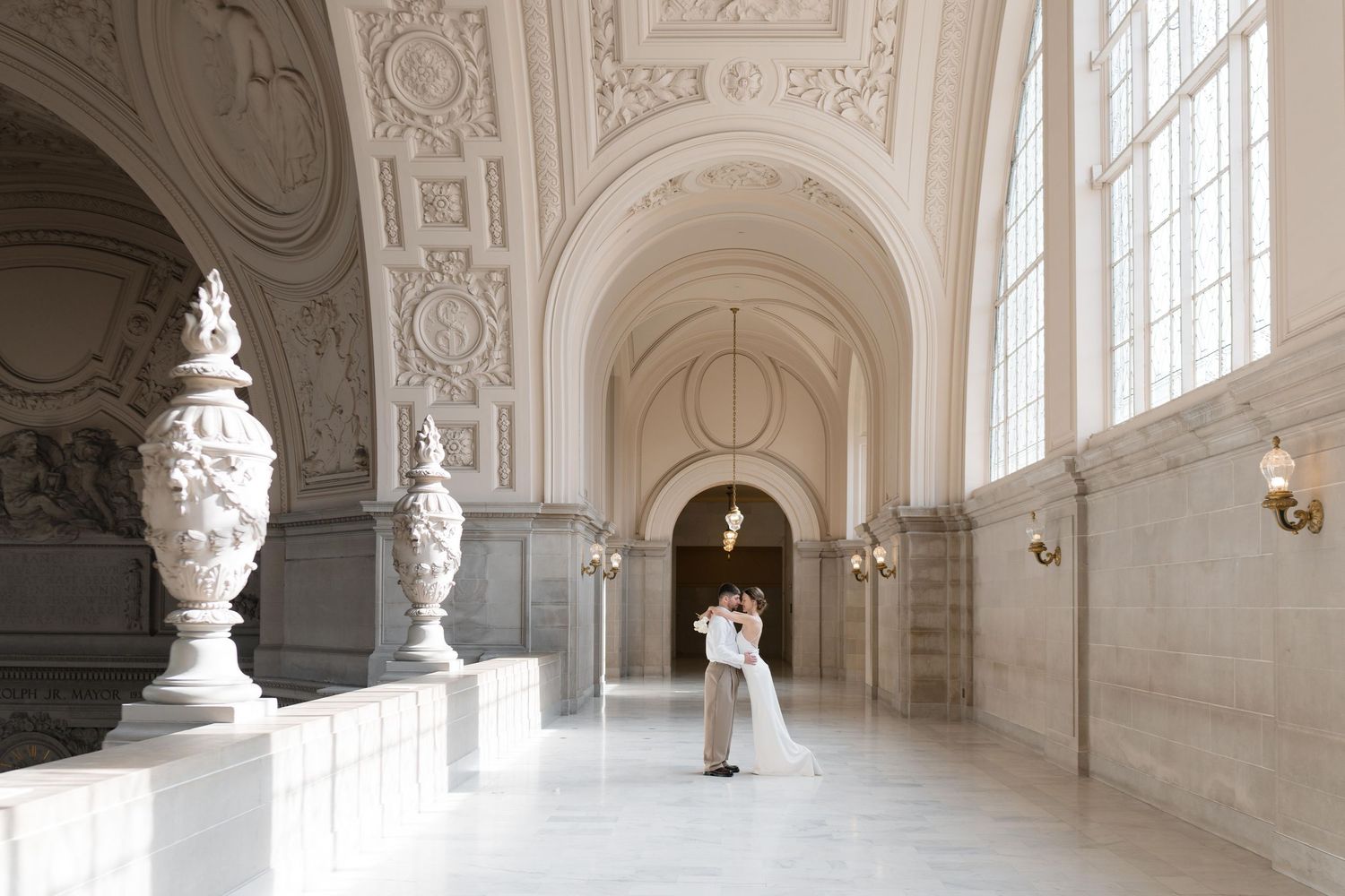 A figure in white stands in an elegant marble hallway with ornate architectural details and decorative urns at San Francisco City Hall.