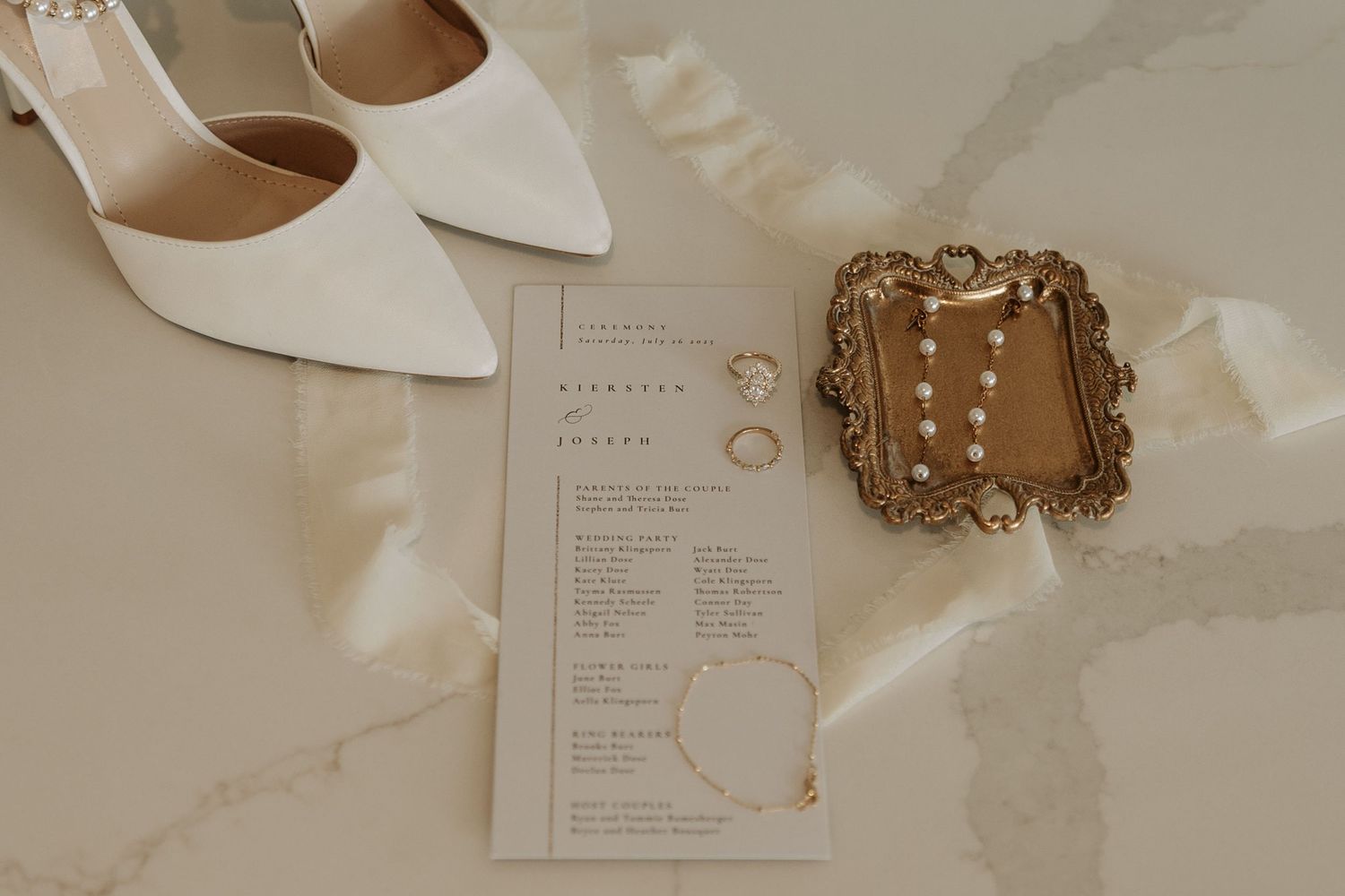 A neutral-toned wedding flat lay featuring invitations, shoes, rings, and delicate decor elements arranged on a marble surface.