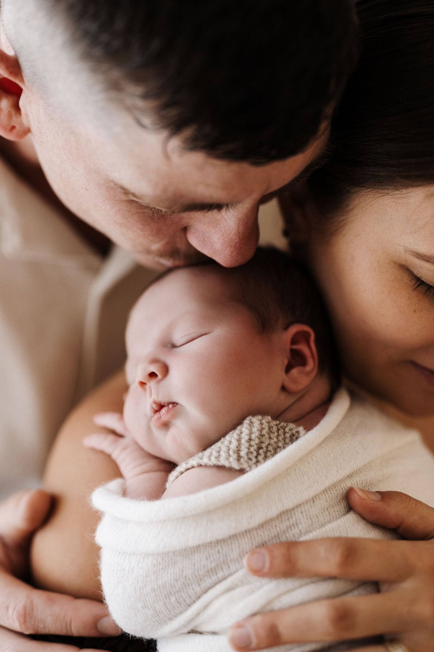 A tender moment between parent and newborn baby wrapped in soft white knit blanket as they share gentle kisses.