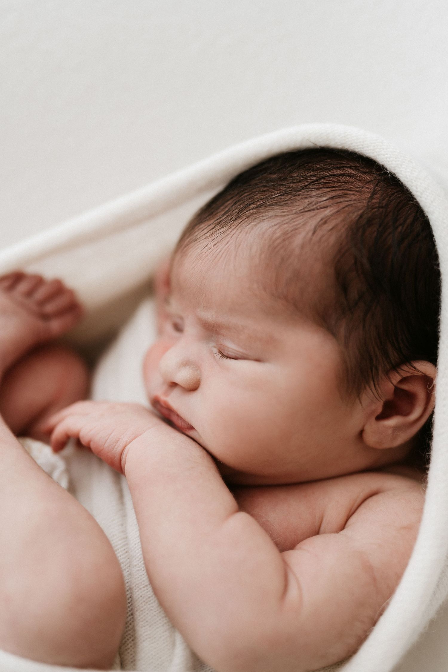 Peaceful sleeping newborn baby wrapped in soft white blanket, photographed in gentle natural light from multiple angles.