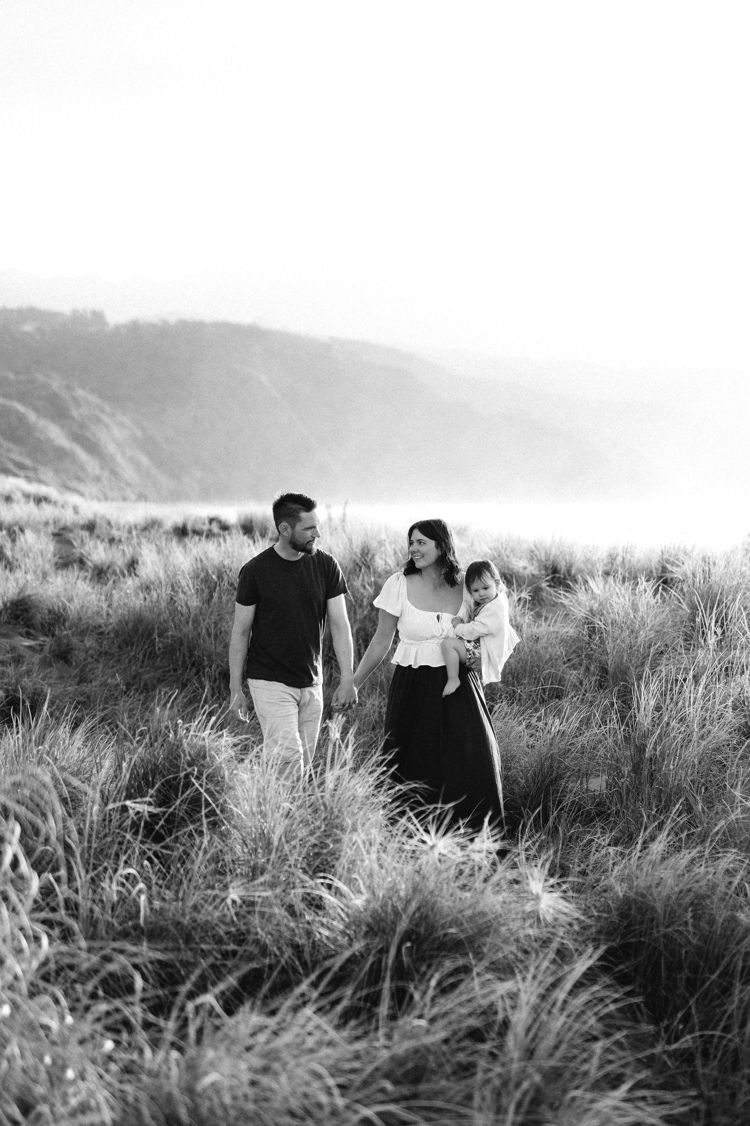 Black and white photo of a family walking through coastal grass, mother holding baby, father beside her, hills in background.