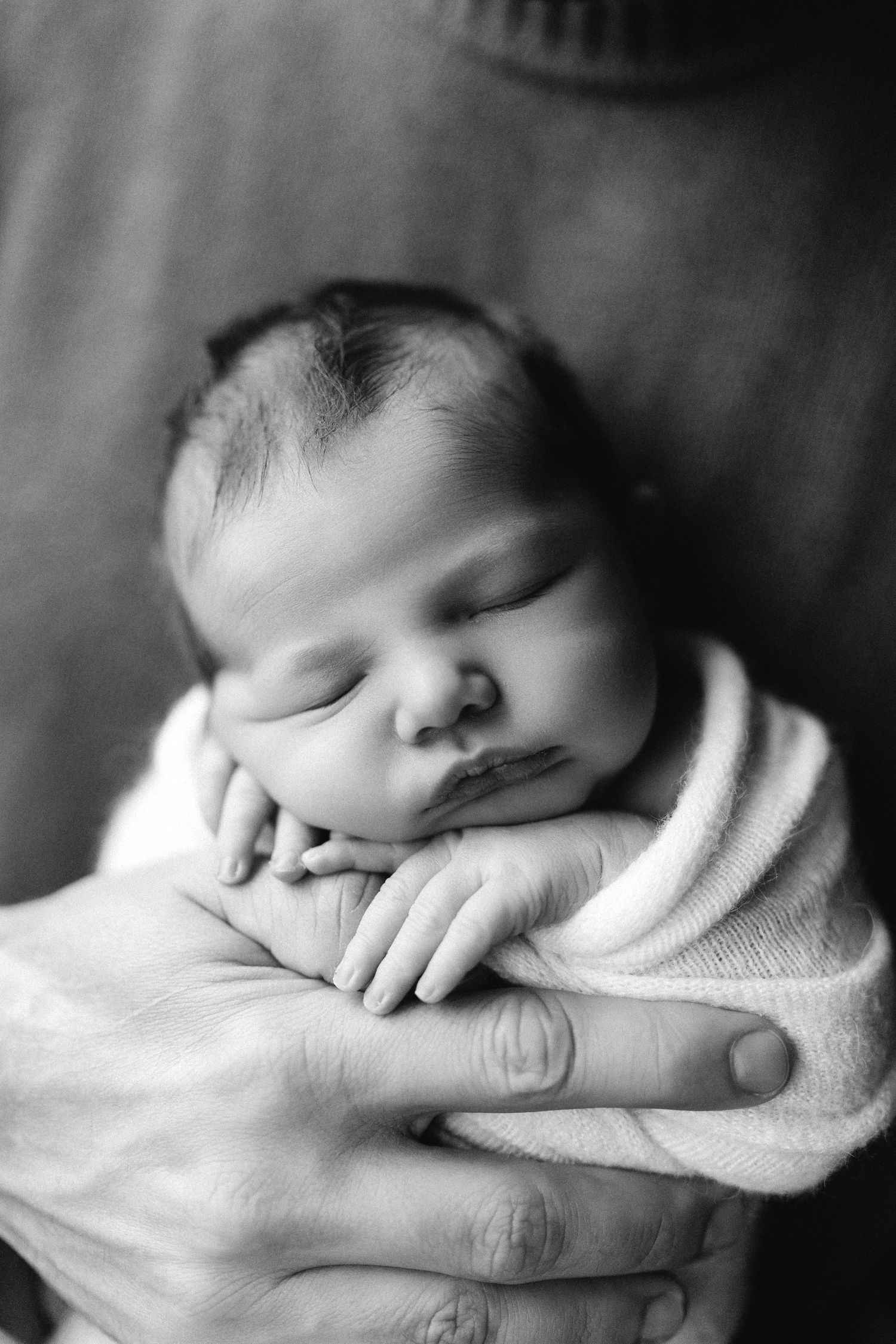 Black and white portrait of a peacefully sleeping newborn baby cradled in caring hands against a soft background.