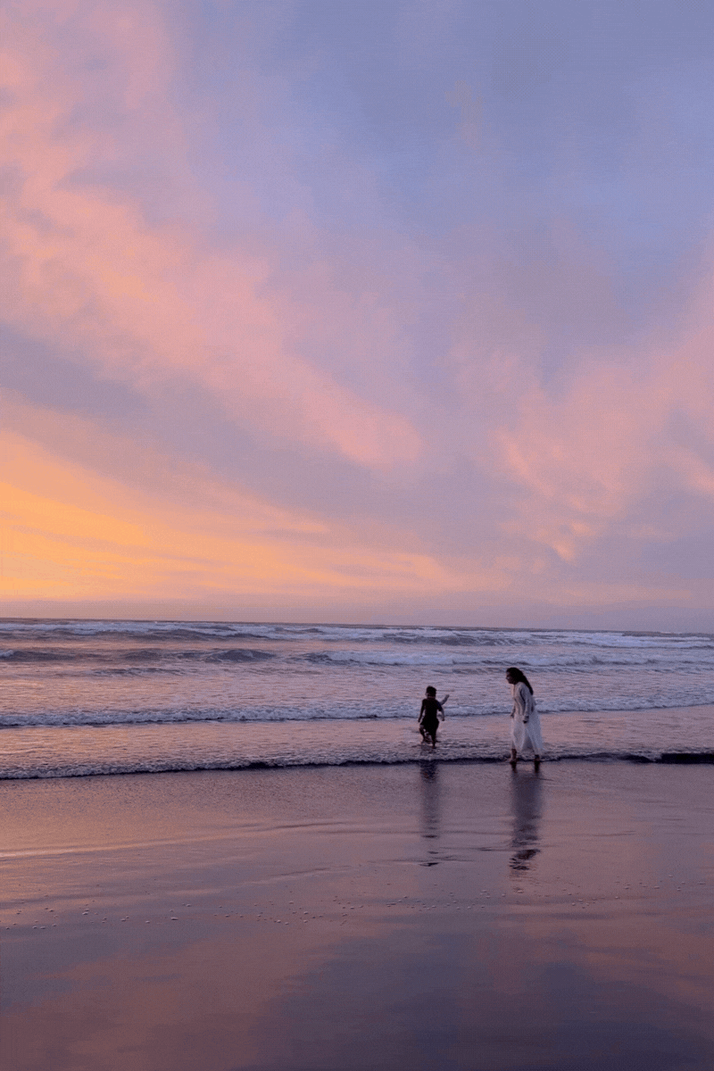 Two silhouettes on a beach at sunset with pink and purple clouds reflecting in the wet sand and ocean waves.