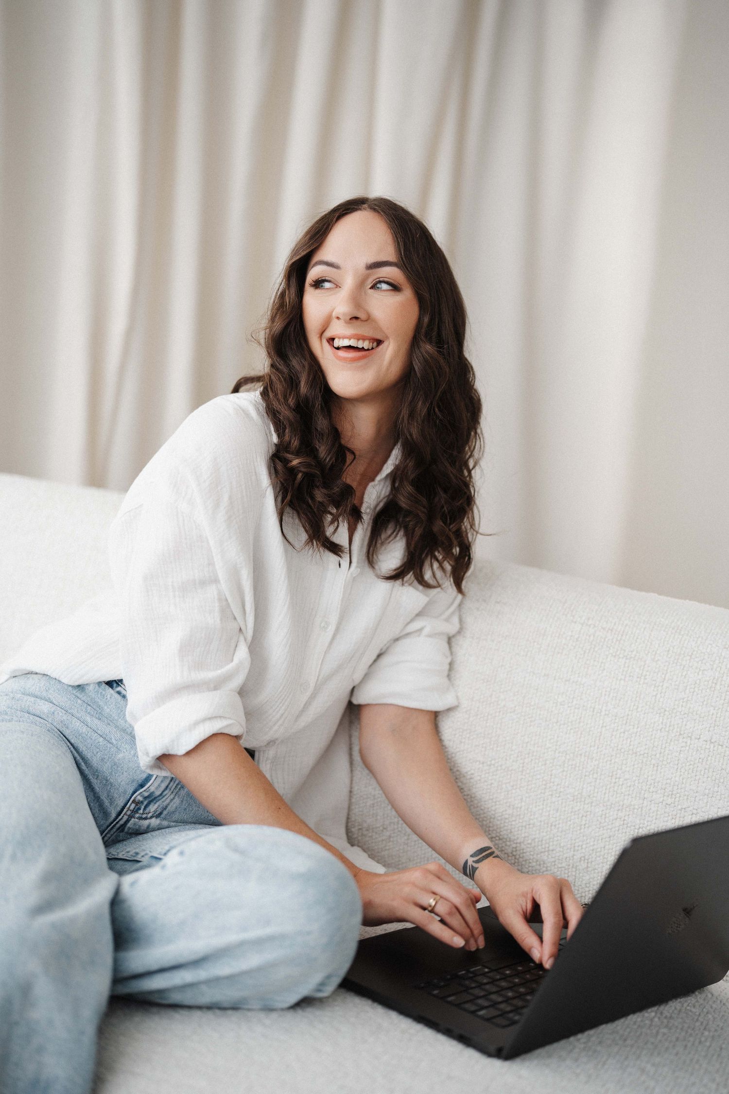 Woman in white shirt and jeans lies on white couch while working on laptop with joyful expression.