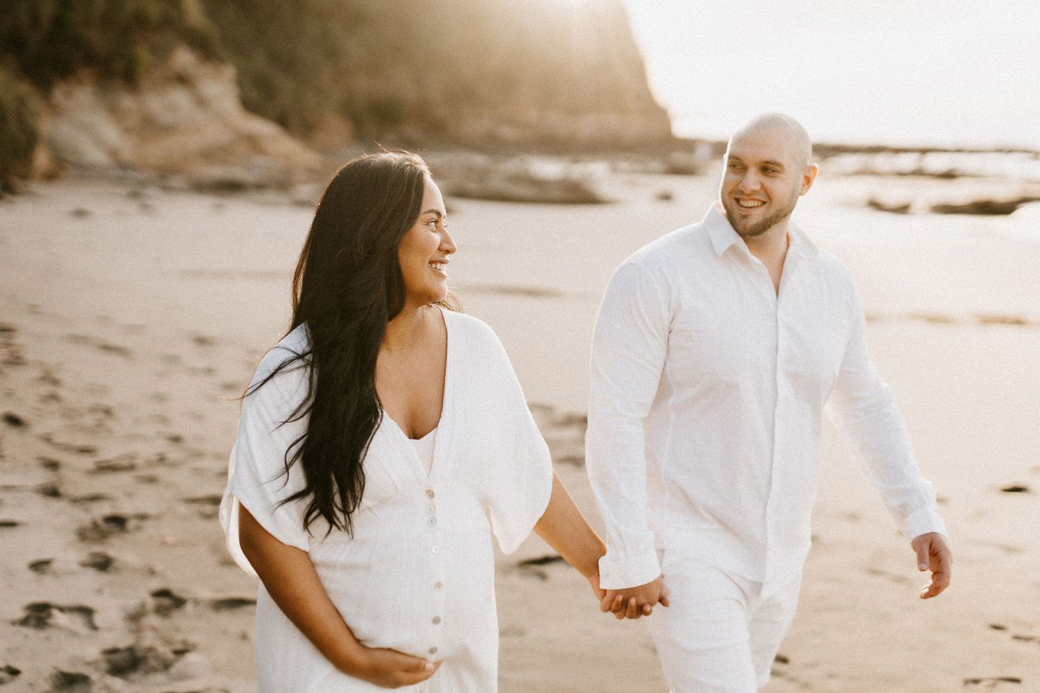 Couple in white outfits walks hand in hand on scenic beach during sunset.