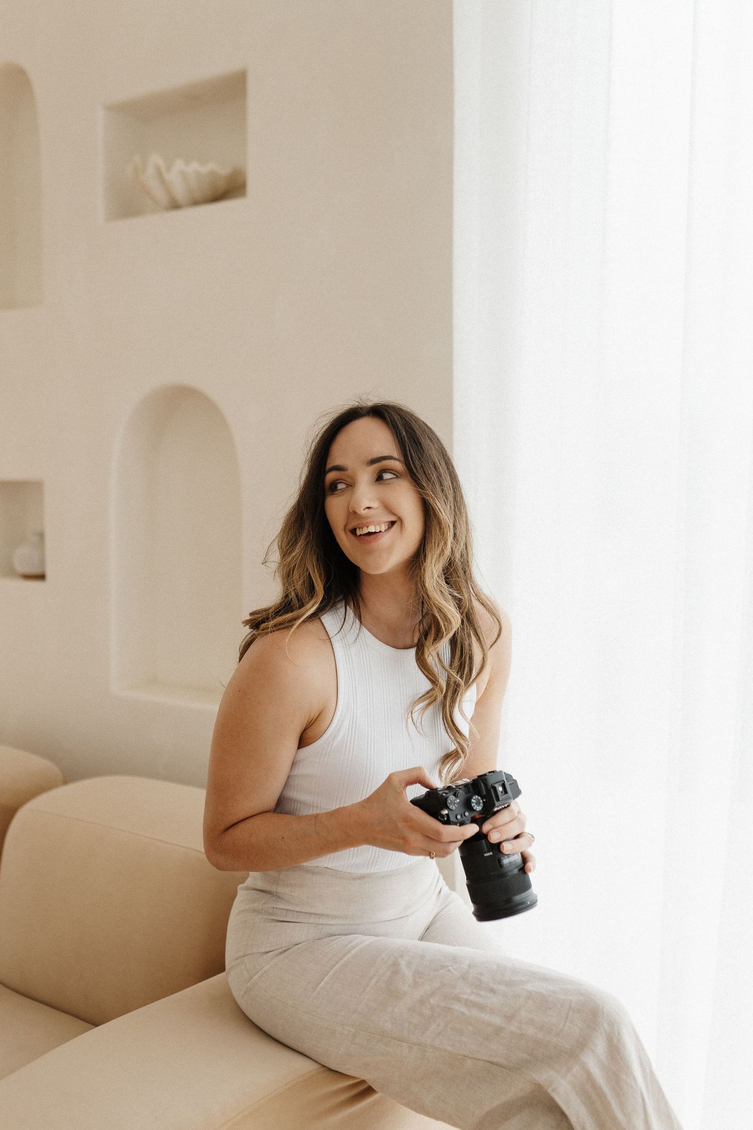 Professional photographer in white outfit sits casually on beige couch holding camera in bright minimal setting.