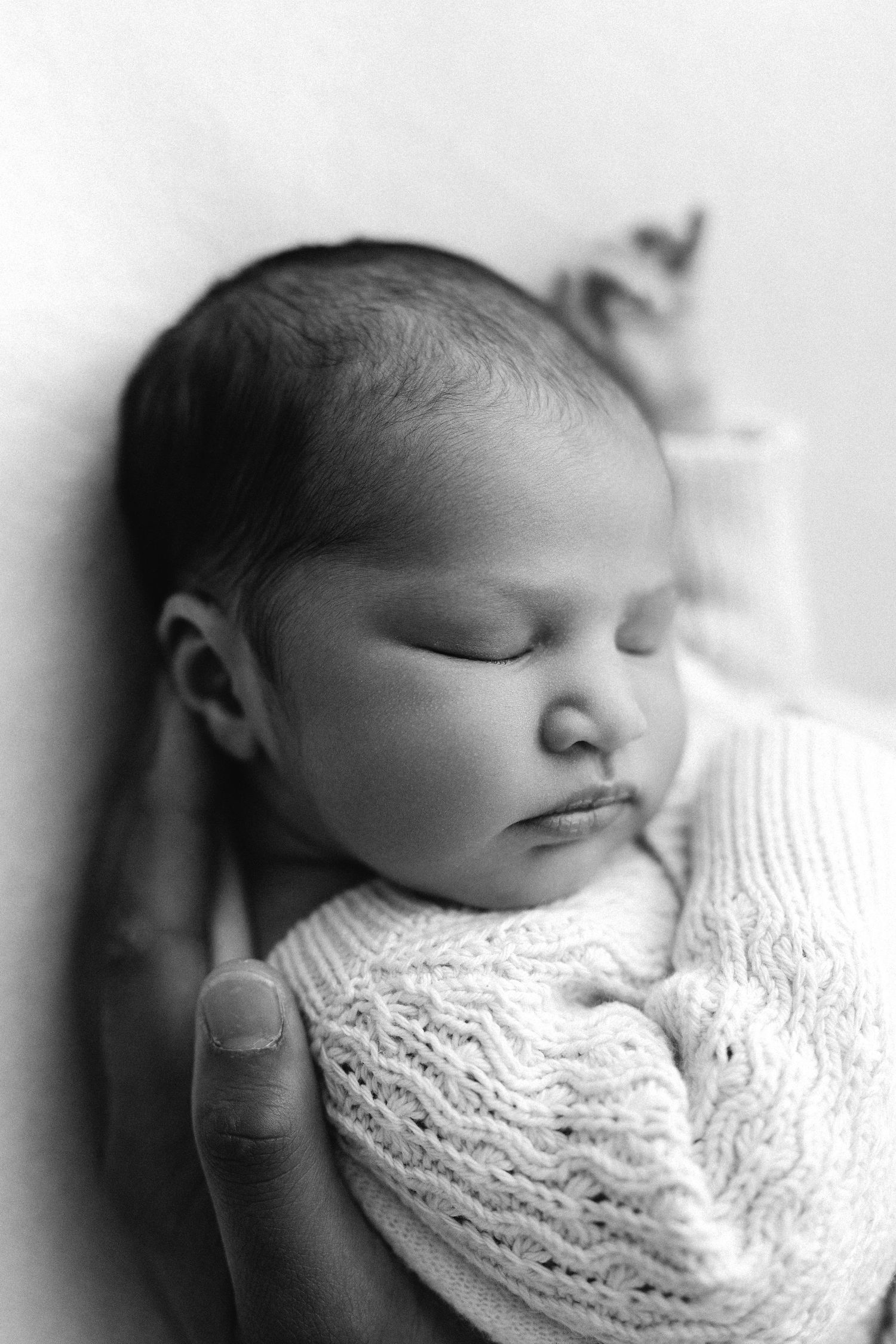 Black and white photo of a sleeping newborn baby wrapped in a knit blanket, cradled in an adults hand.