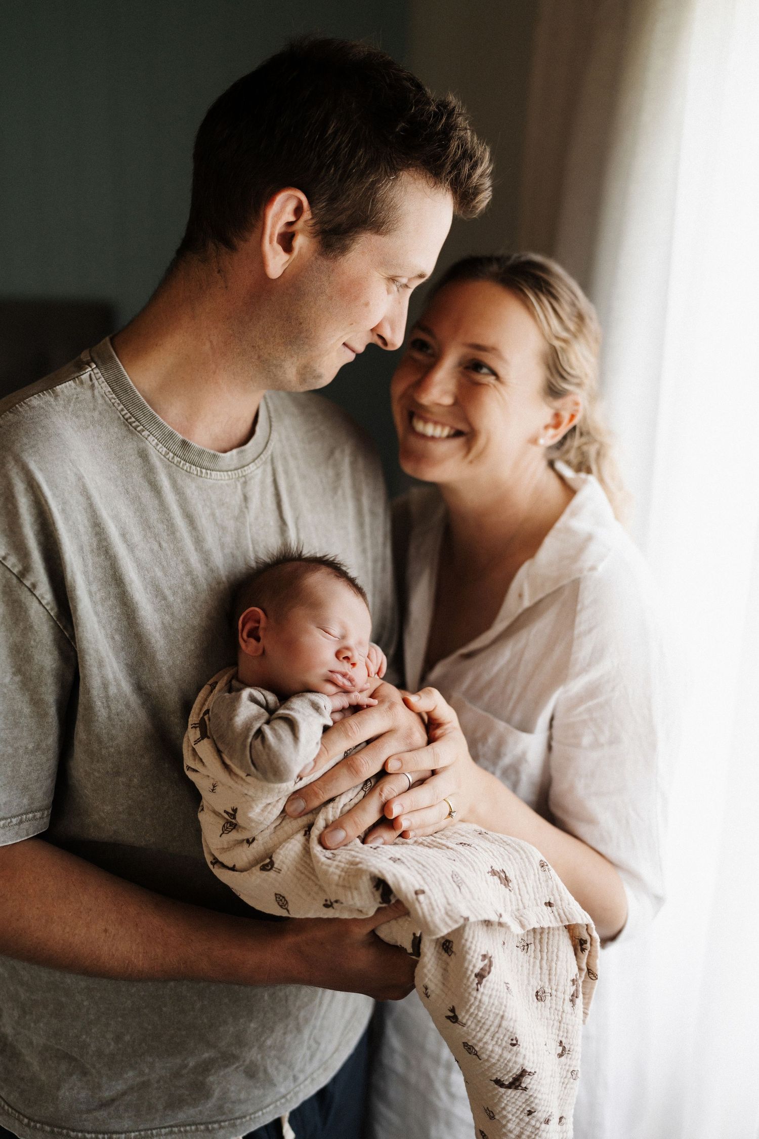 Loving parents hold their sleeping newborn baby by a window in their home during a tender family moment.