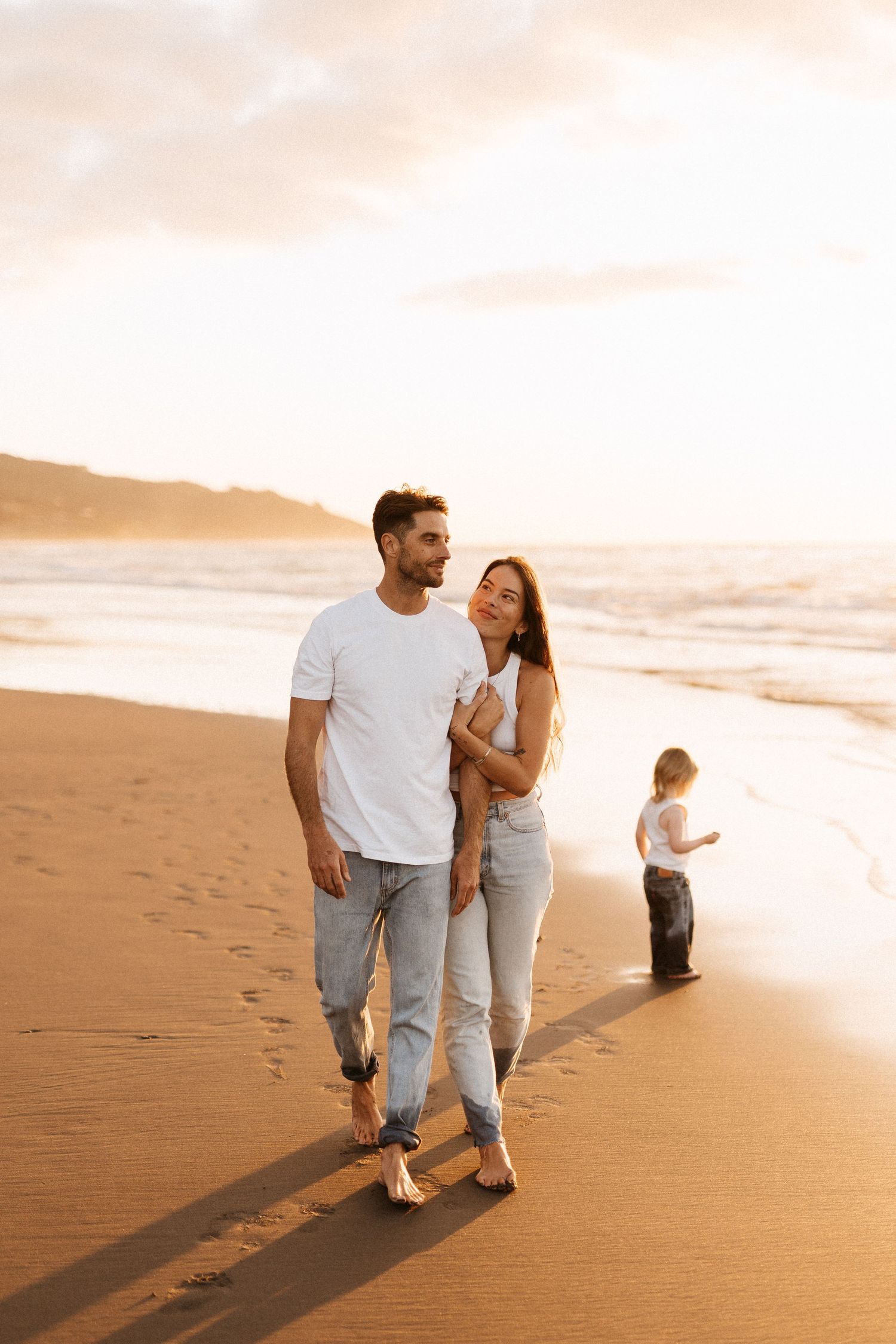 Couple walking barefoot on sandy beach at sunset while child plays near ocean.