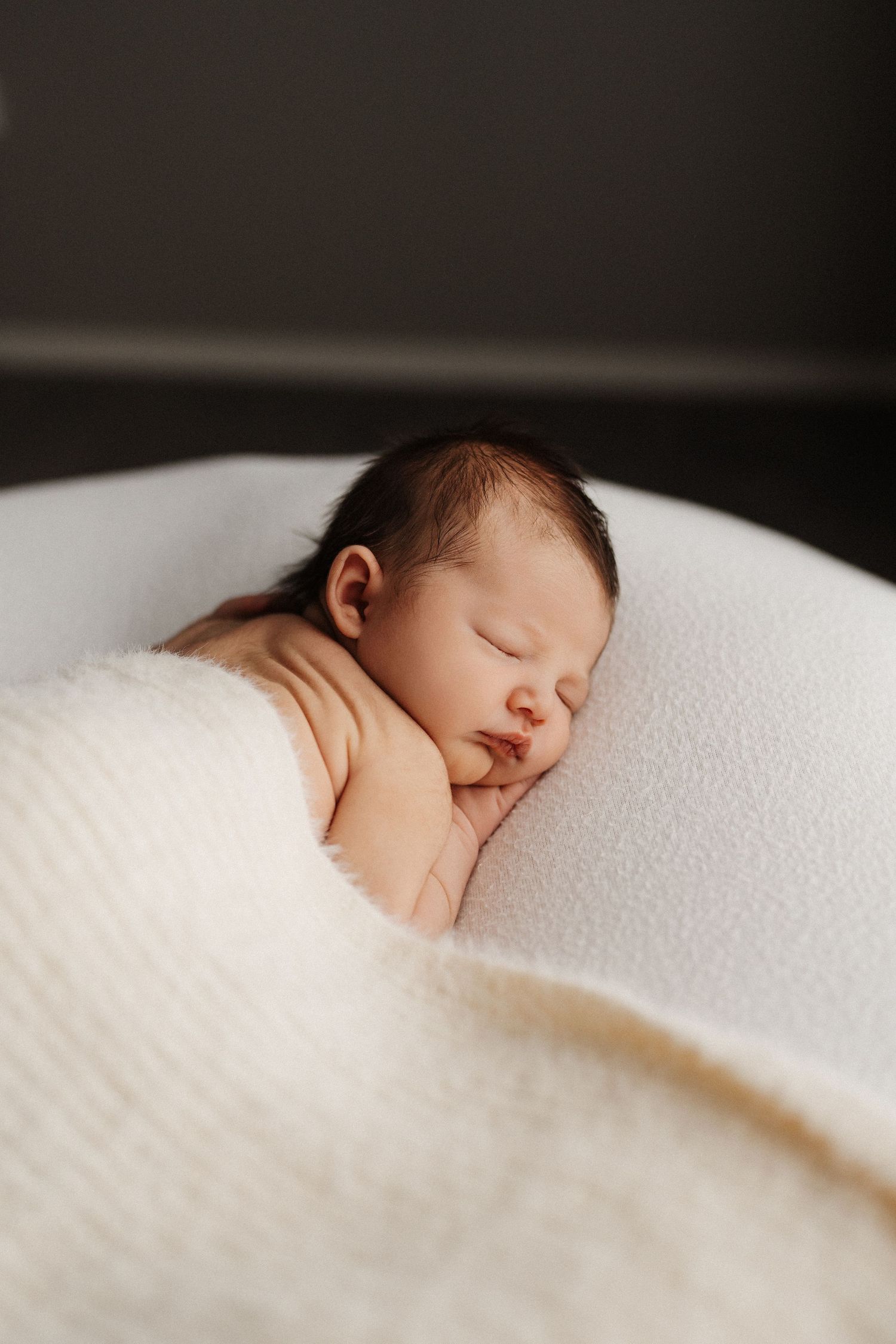 A sleeping newborn baby rests peacefully on soft white bedding in natural lighting.