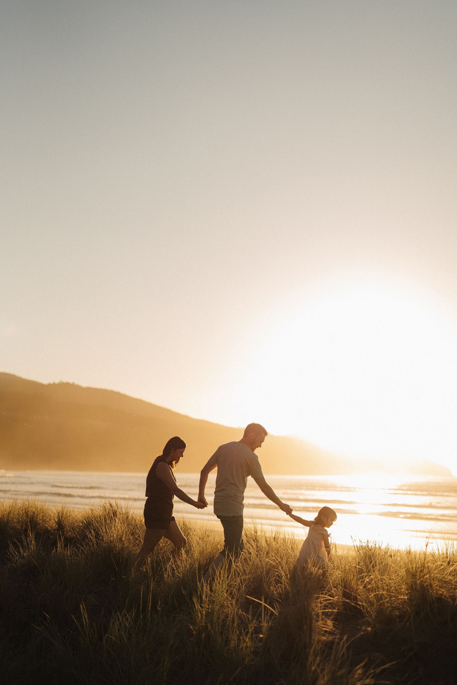 Silhouettes of people walking through tall grass on a beach at sunset with golden light streaming across the landscape.