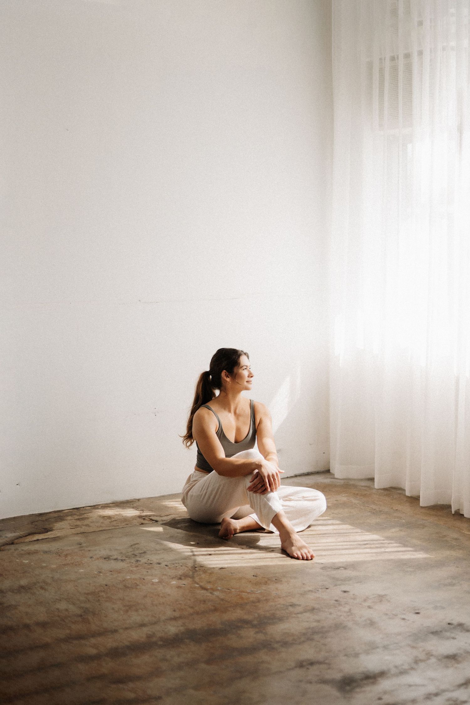 A person in light-colored clothing sits peacefully on the floor near a sunlit window in a minimalist room.