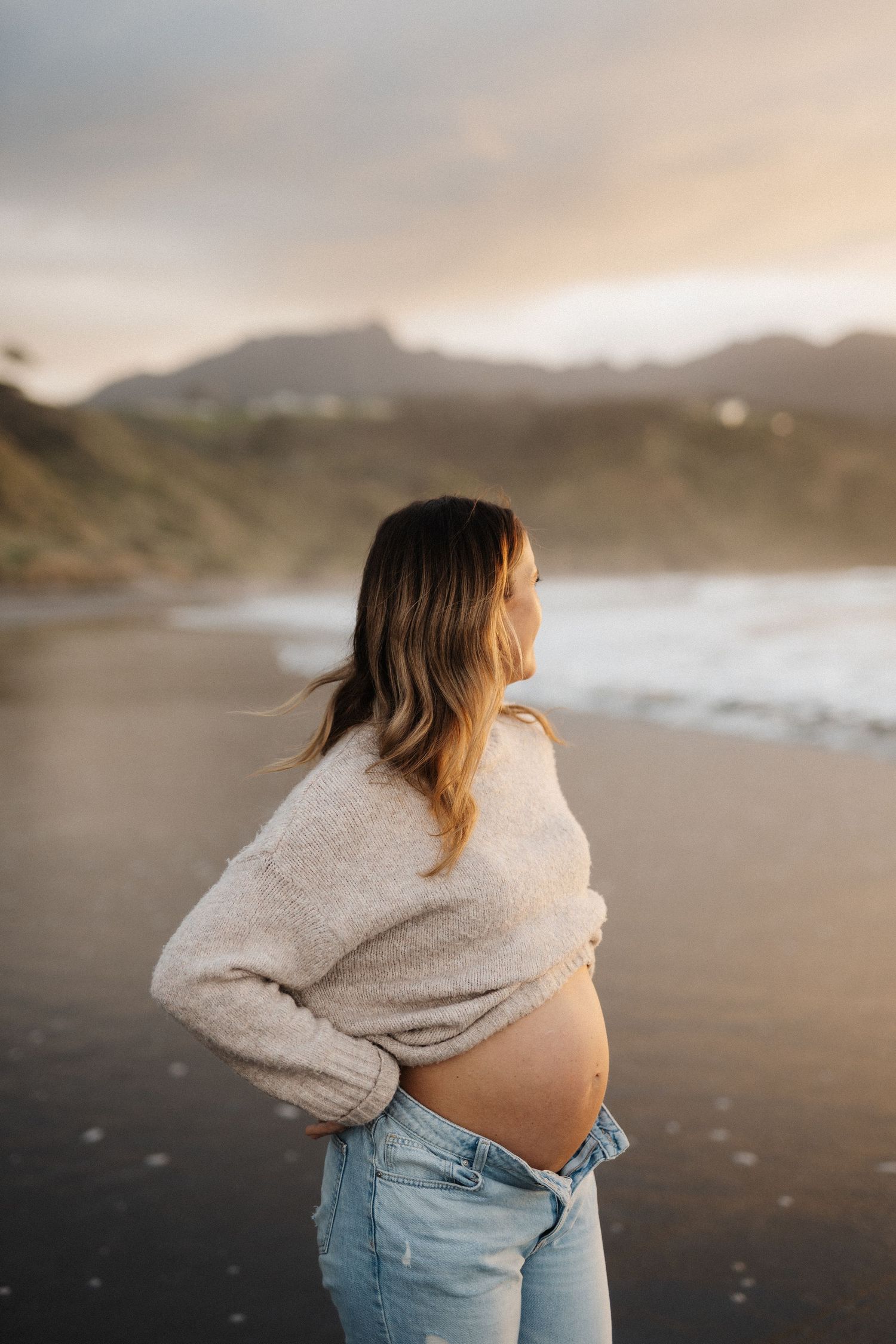 Person in cozy sweater and jeans stands on misty beach at sunset with mountains in background.