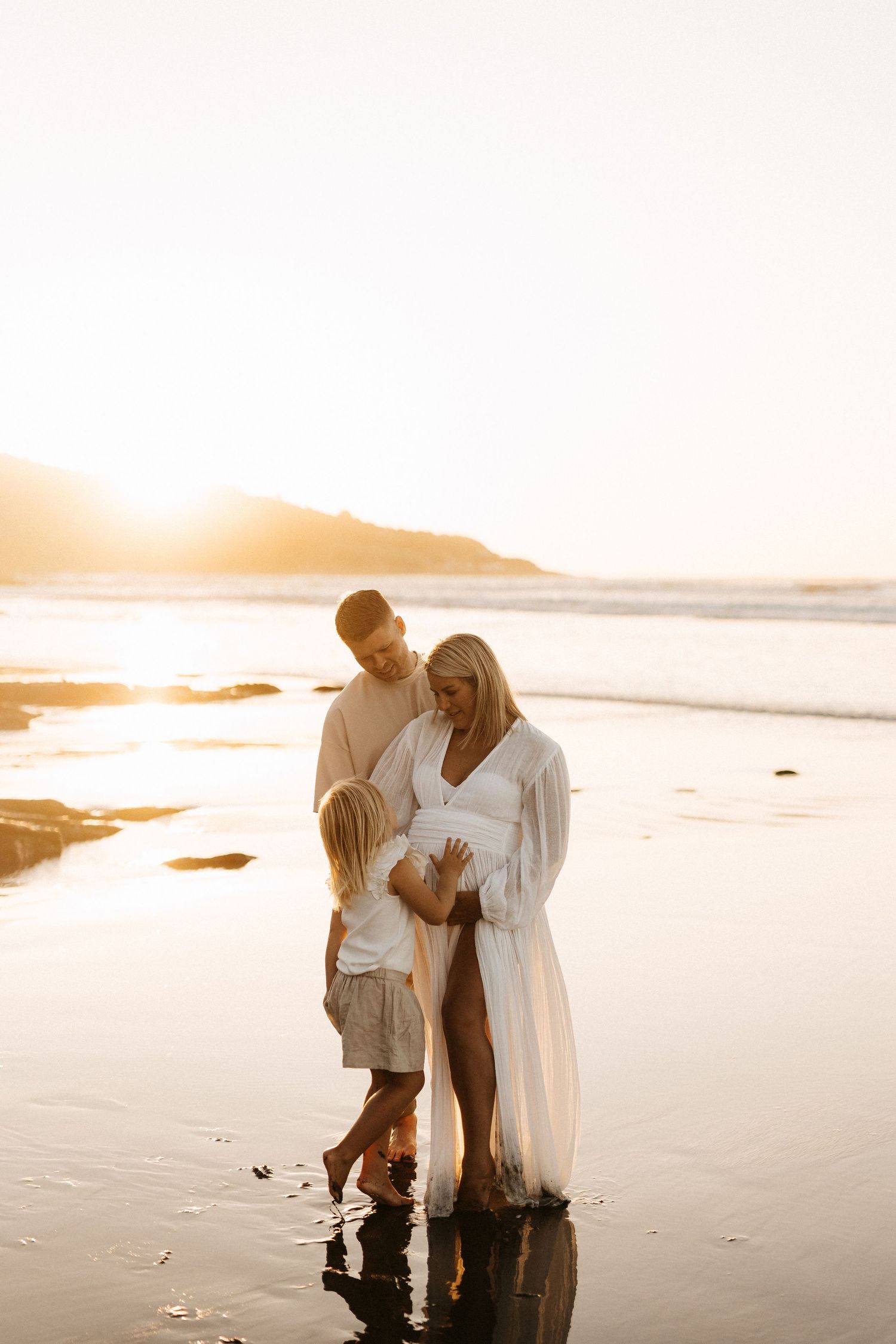Family in white clothing stands together on wet beach sand at sunset.