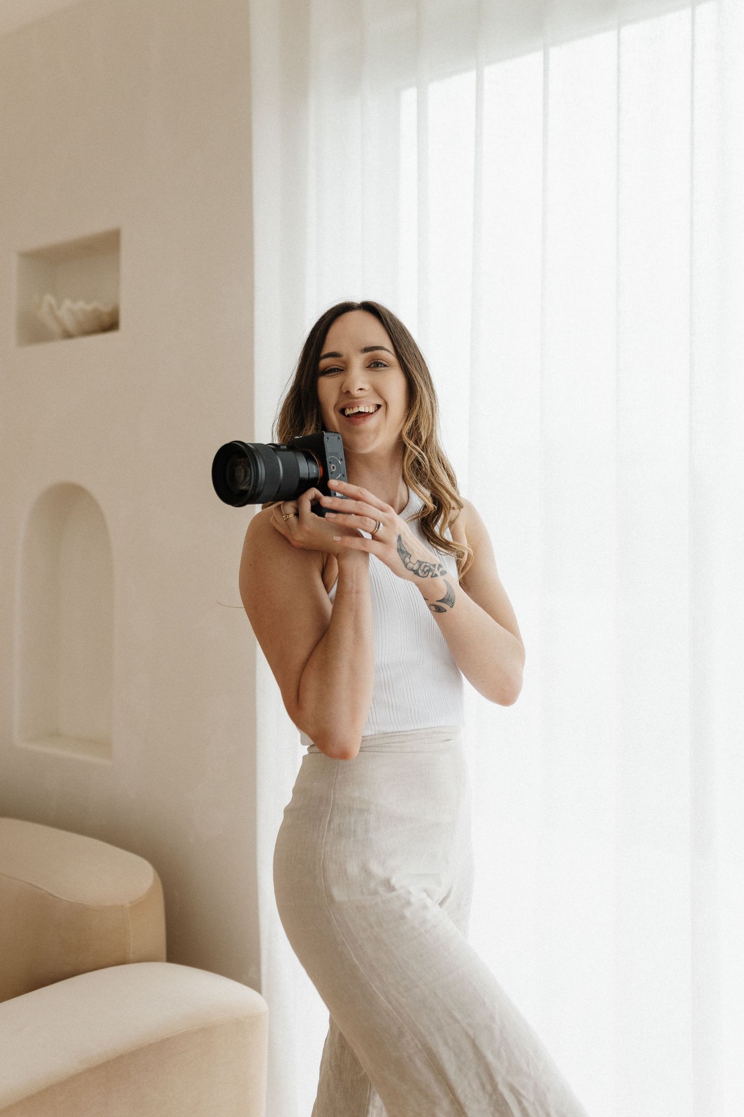 Elegant photographer in white dress takes a self-portrait in a bright minimalist room using professional camera.