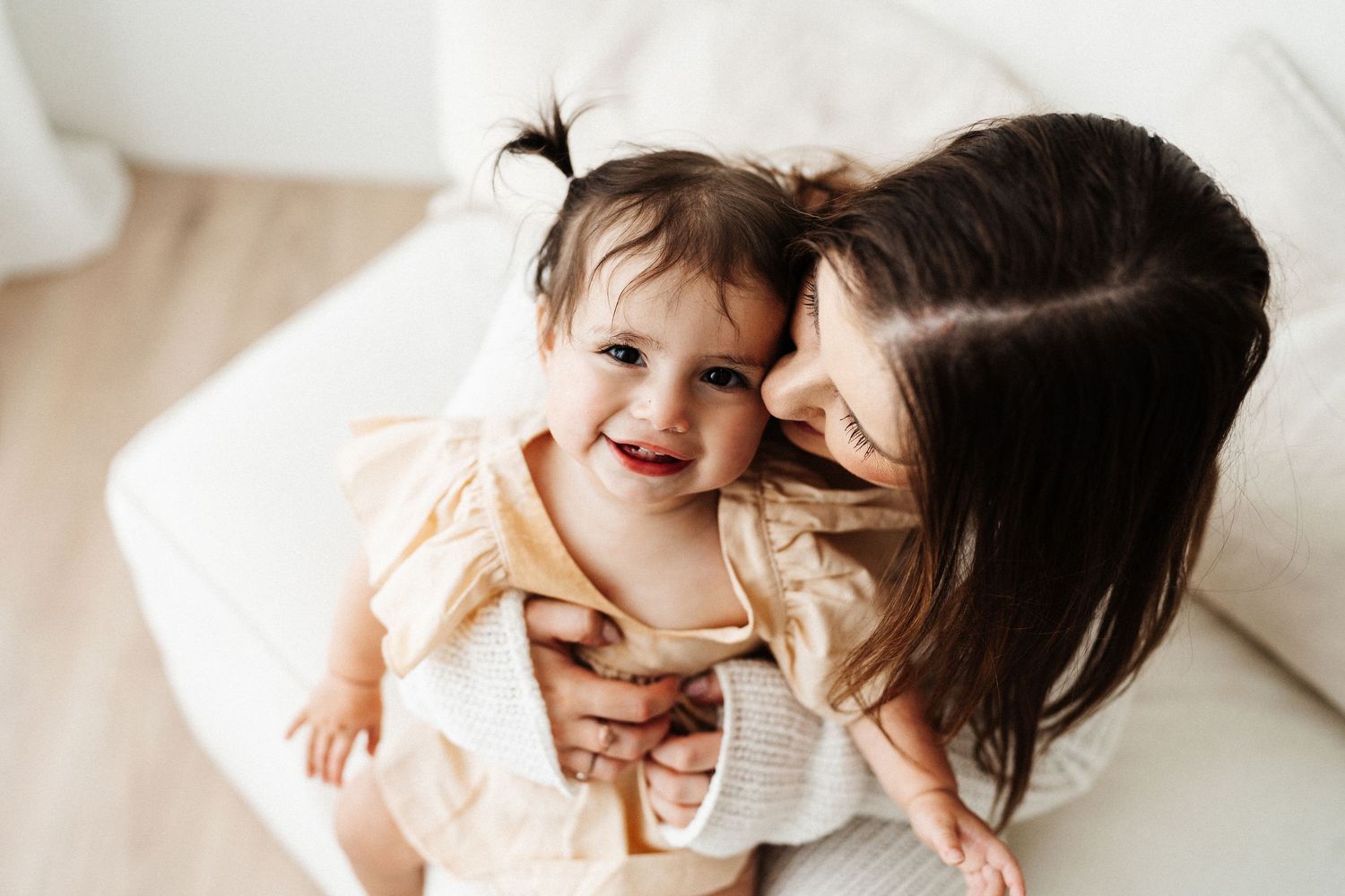A caring moment between parent and small child laughing together while sitting on white bedding.