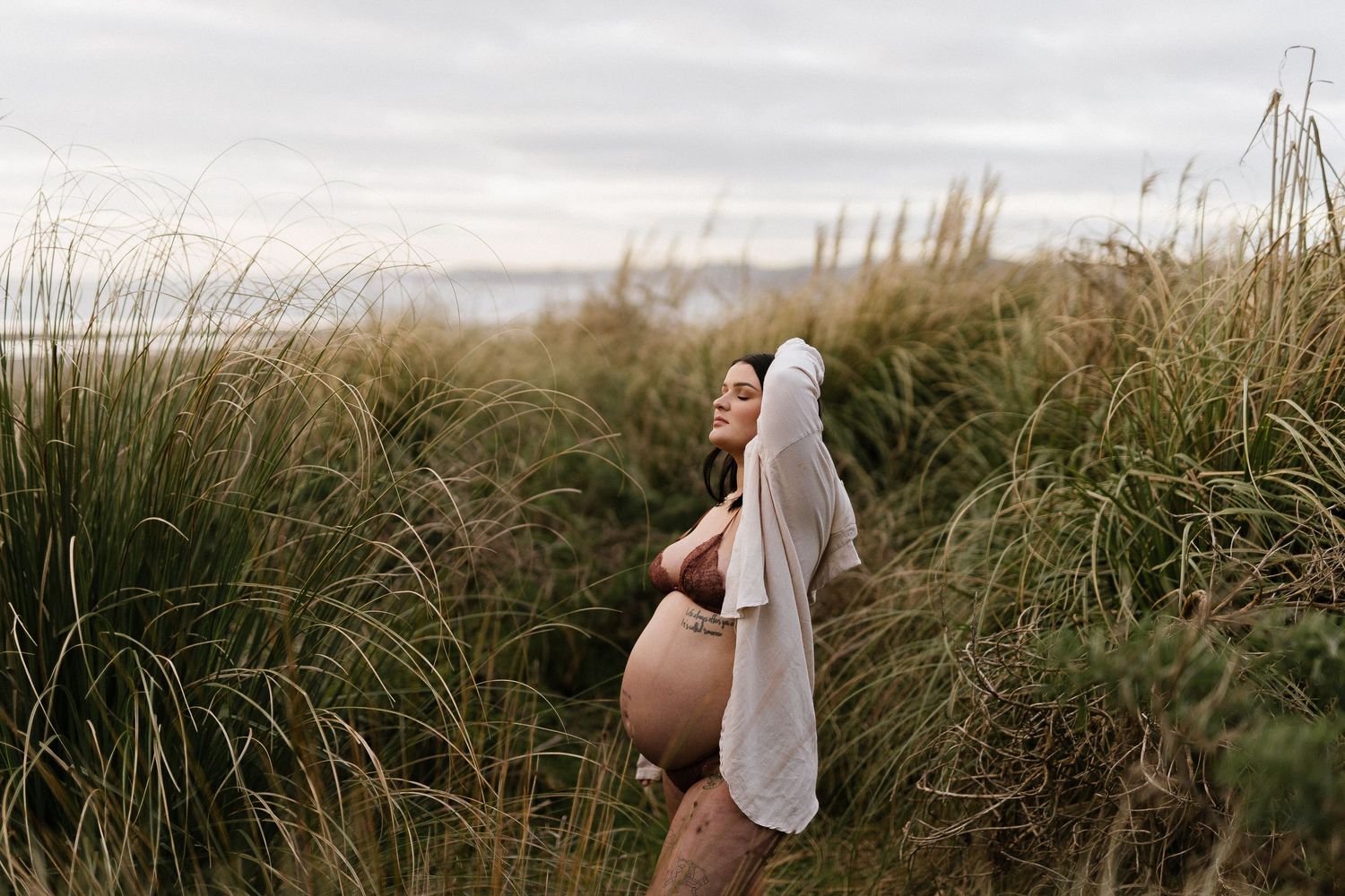 Pregnant woman in white dress poses among tall beach grass at dusk.