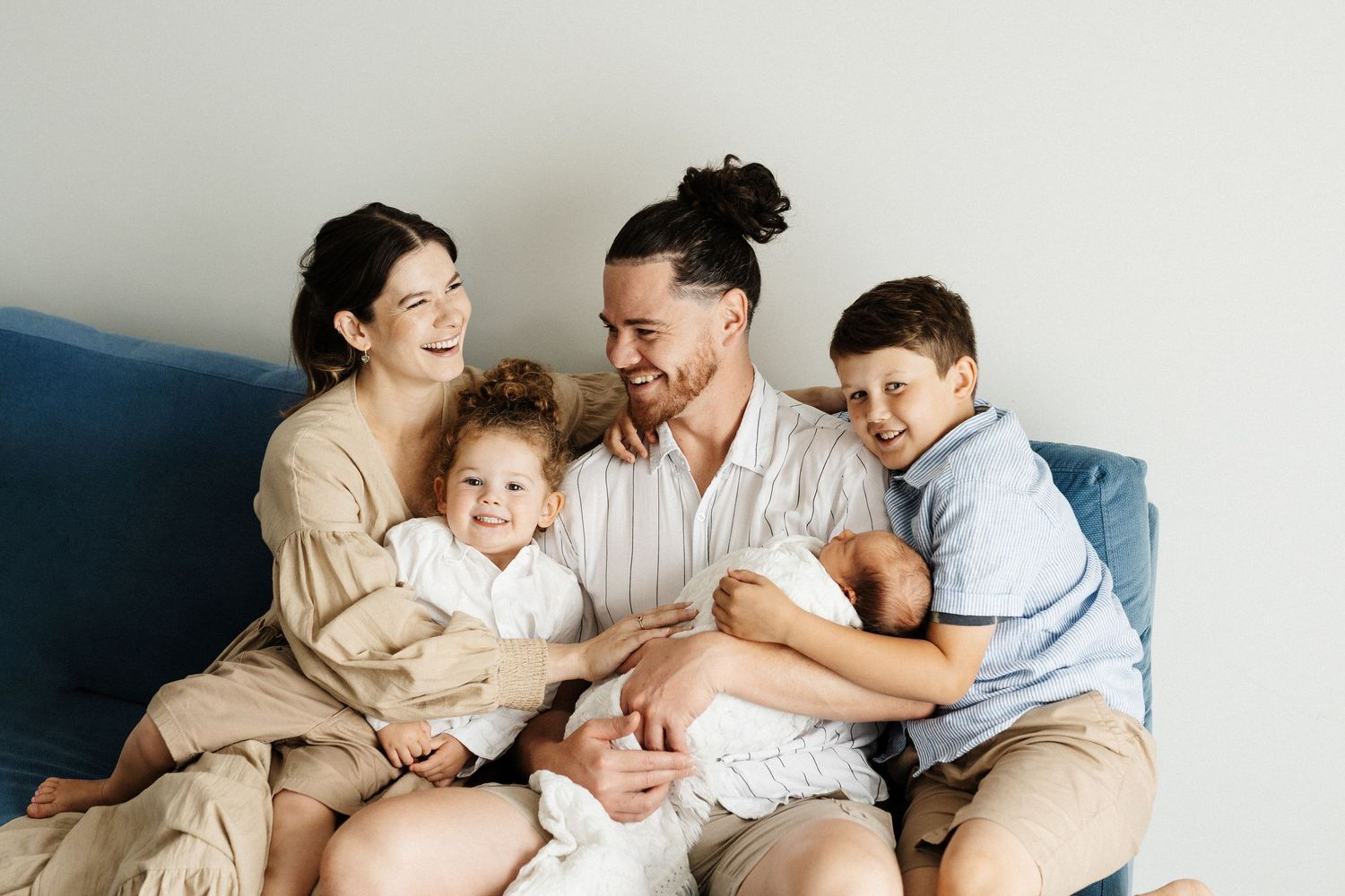 A family of four cuddles together while sharing intimate moments on a white bed.
