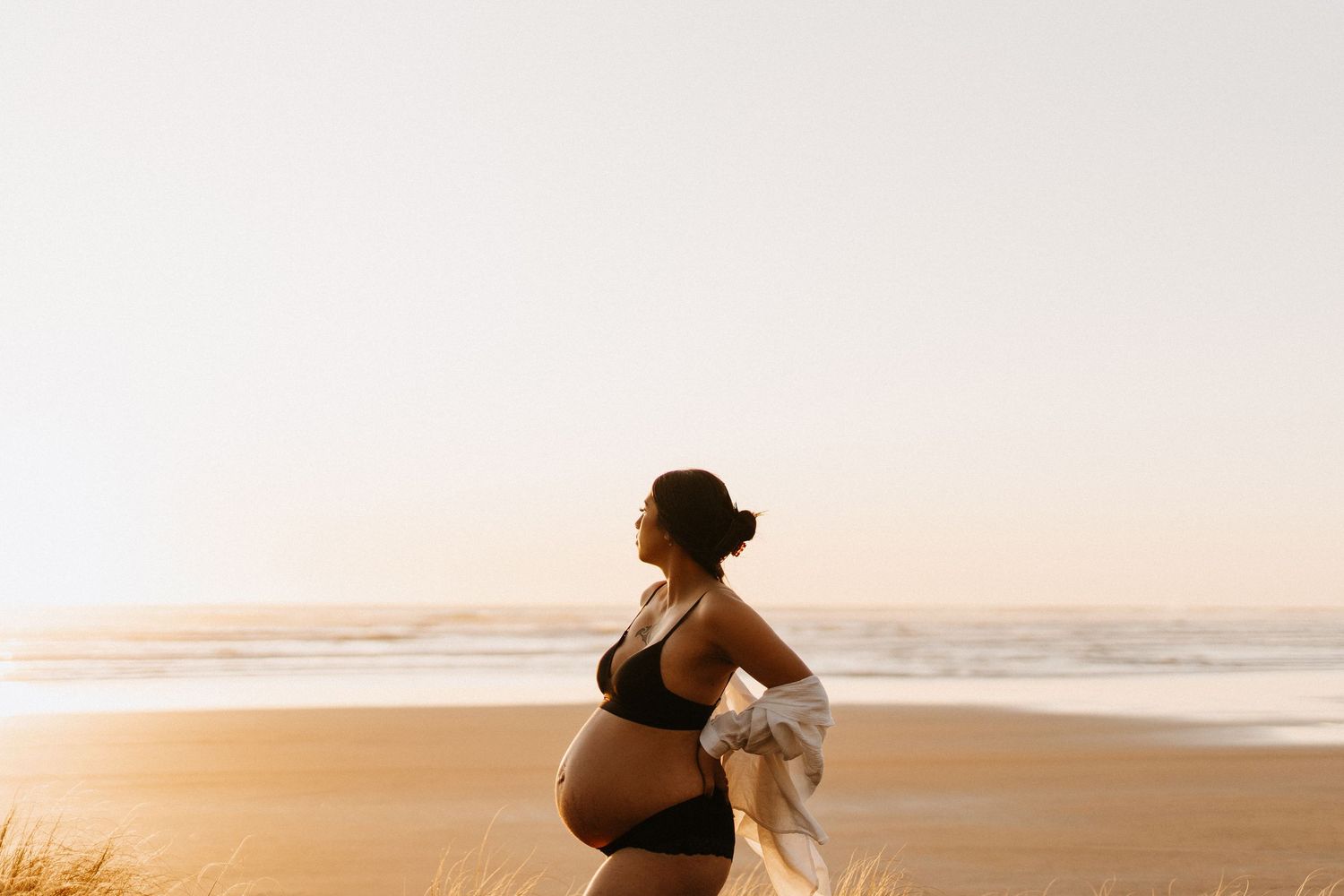 Expectant couple embraces on a scenic beach at sunset during a romantic maternity photoshoot.