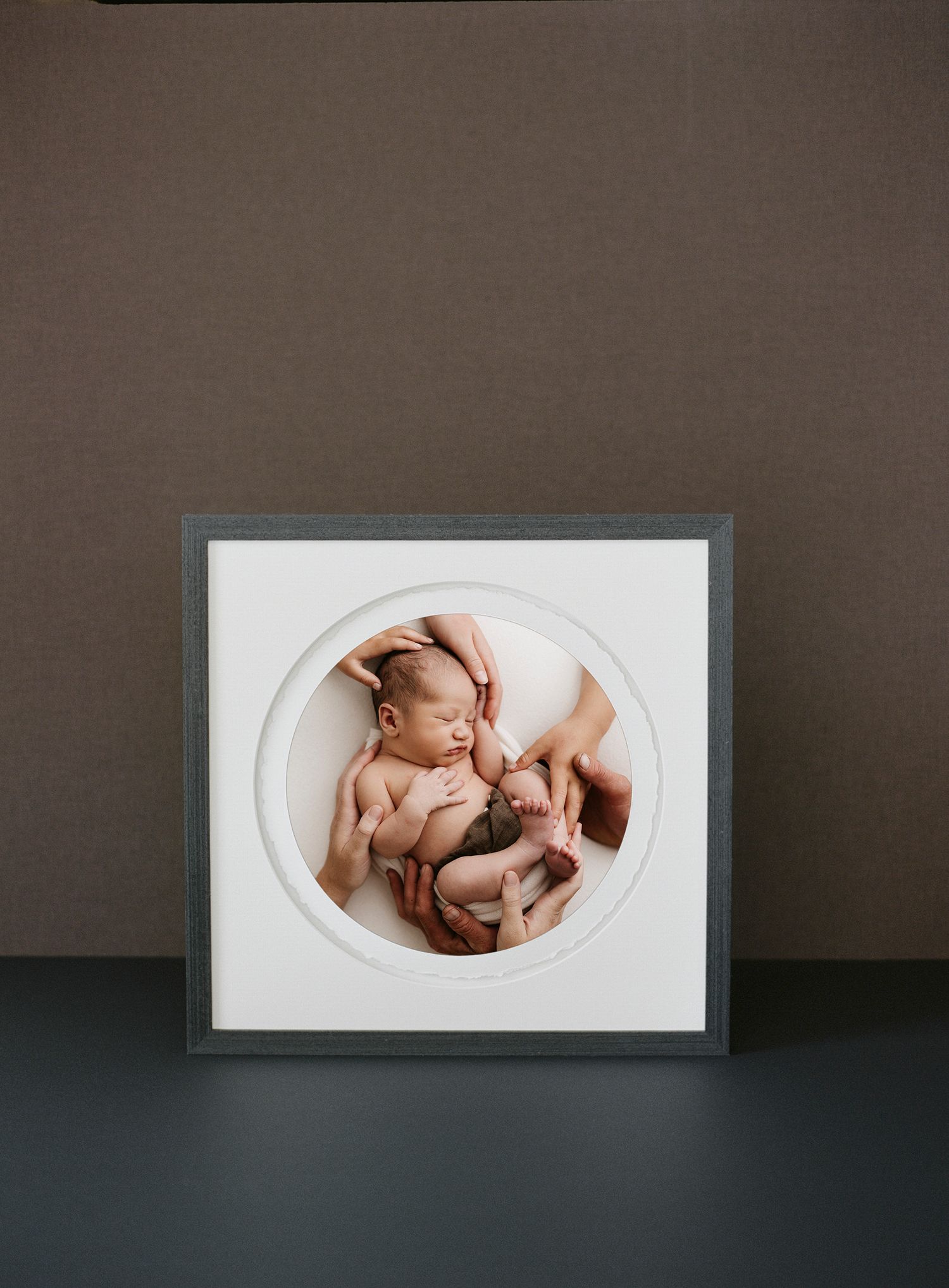 Four minimalist photo frames on a dark wall show artistic newborn baby portraits in oval-shaped mats.