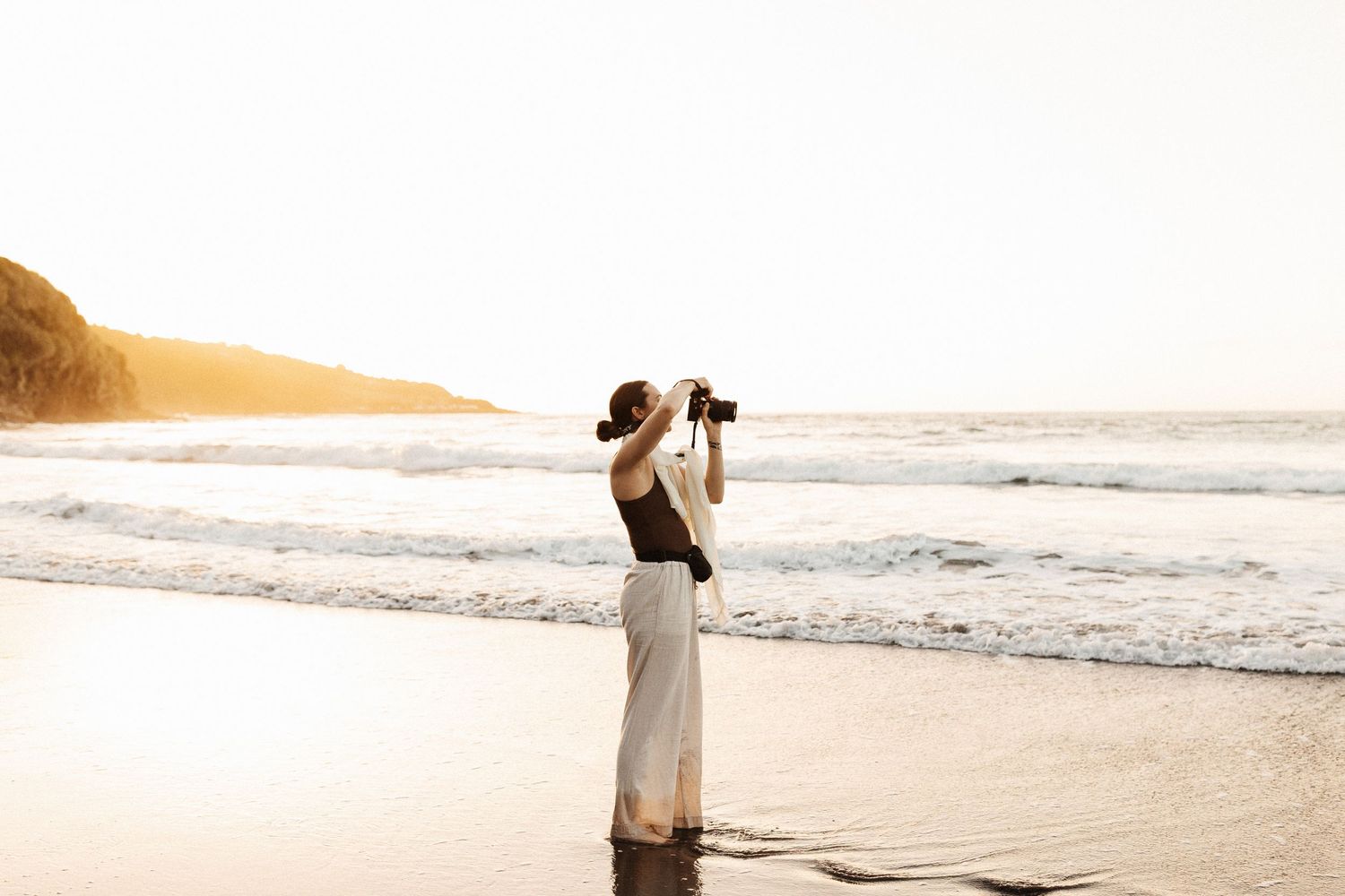 A photographer silhouetted against golden sunset light photographs on a beach.