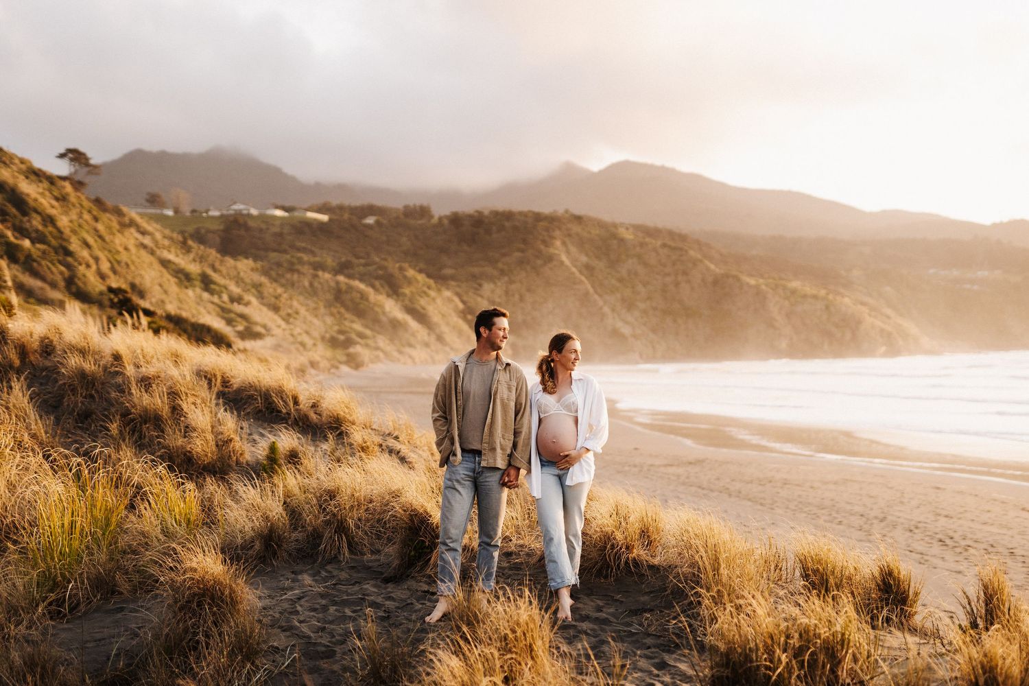 Couple walking along scenic coastal cliffs at golden hour with ocean views and tall grass in the foreground.