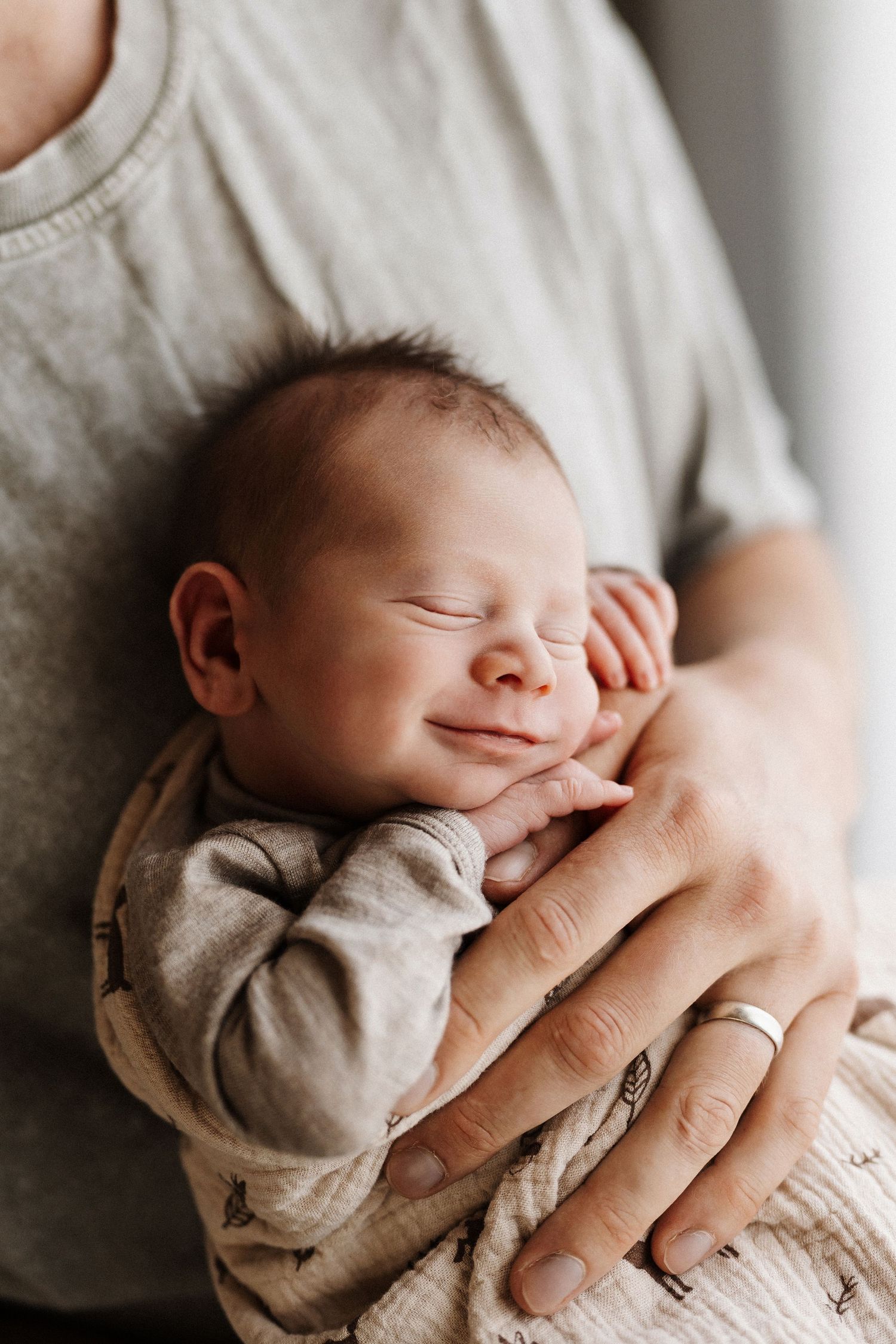 Newborn baby sleeping peacefully in parents' arms while wearing a gray patterned onesie.