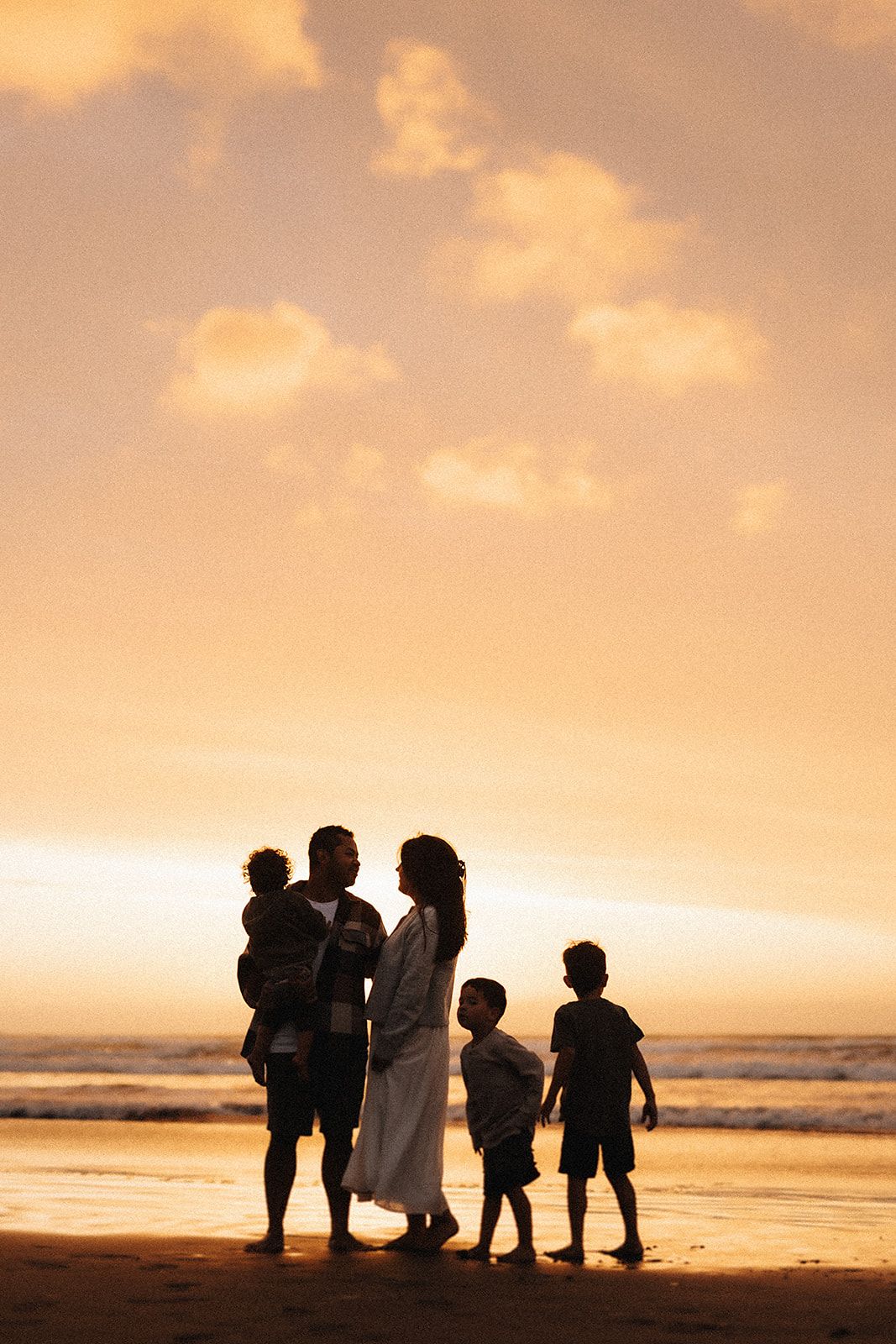 Silhouetted family standing together on beach at sunset with golden sky and ocean waves in background.