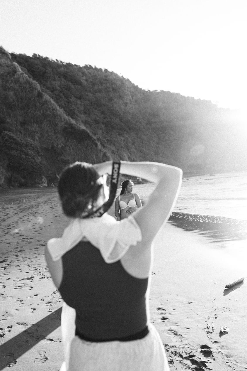 A photographer takes a picture on a sandy beach at sunset with hills in the background.