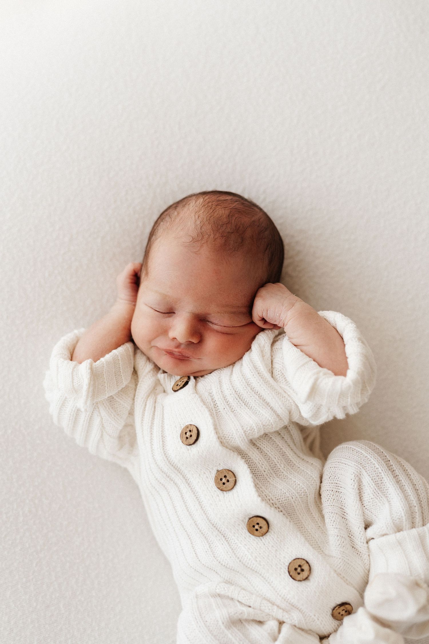Newborn baby sleeps peacefully in white knit cardigan with wooden buttons during professional photoshoot.