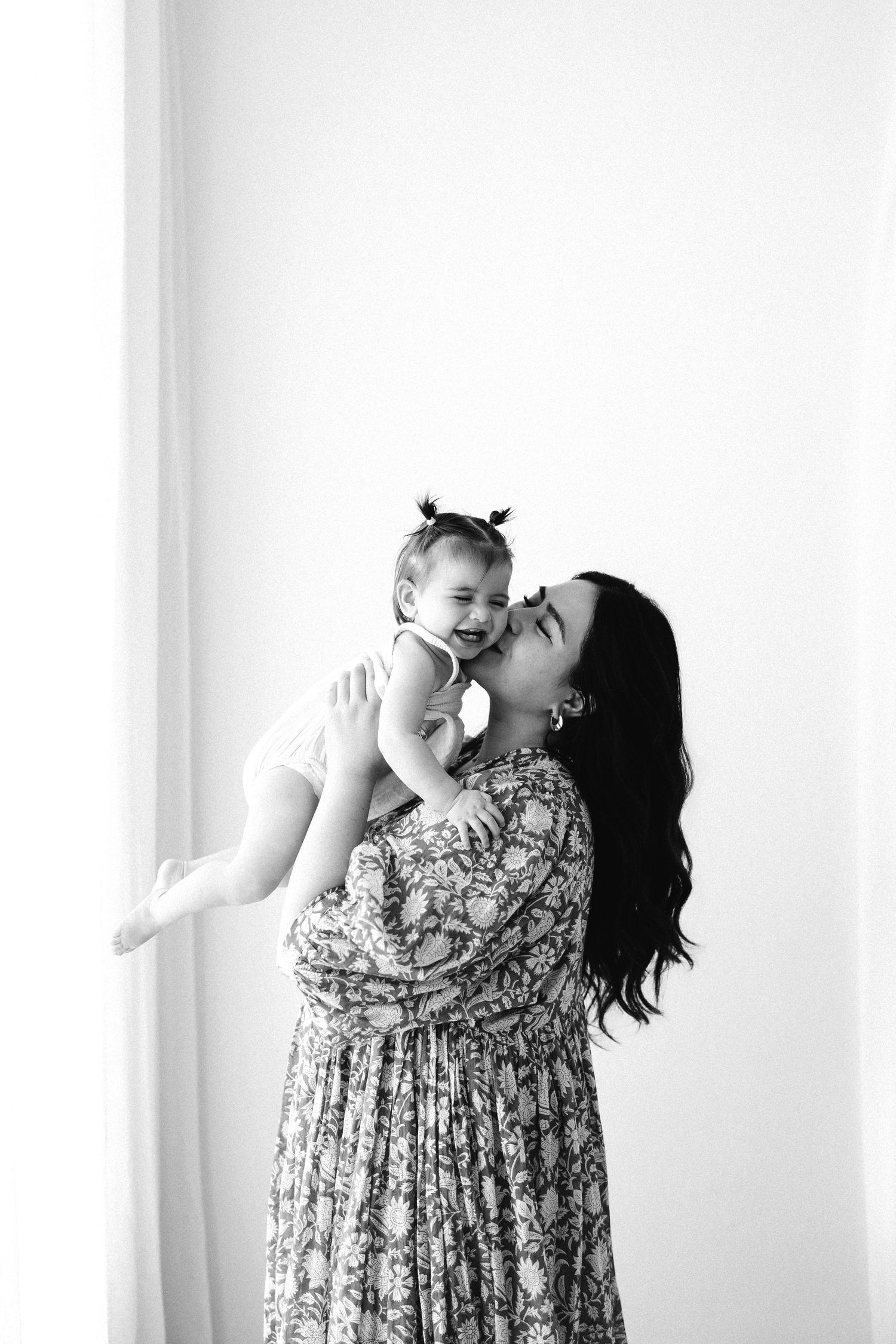 A mother in a floral dress lifts and embraces a joyful baby in black and white triptych photos against a white background.