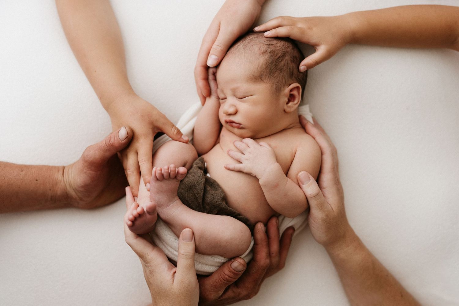 Hands gently hold and cradle a newborn baby in a warm neutral-toned setting during a lifestyle photography session.