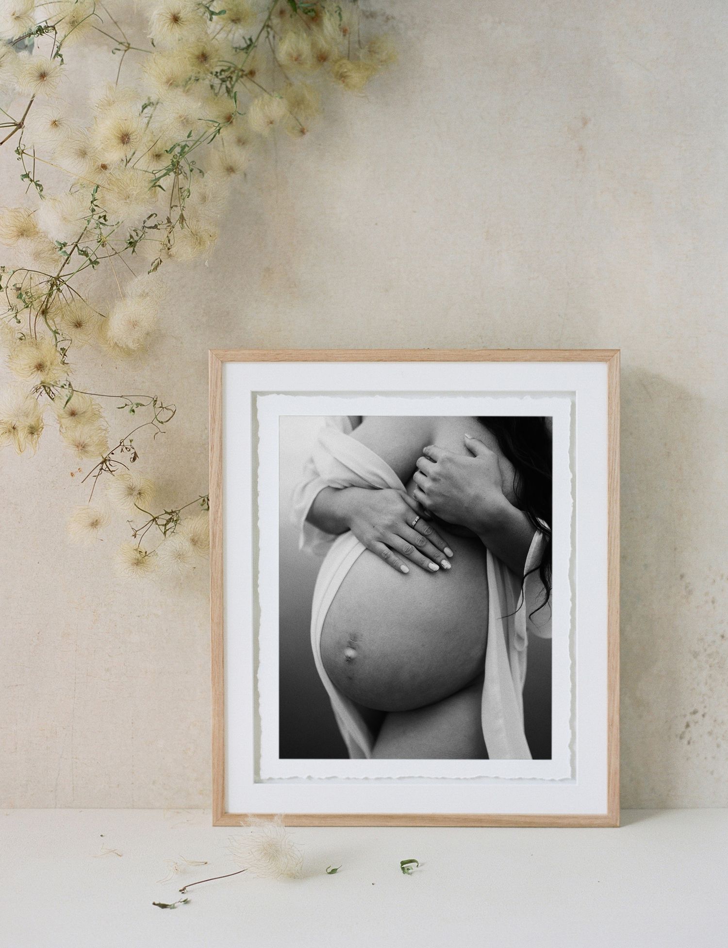 Three black and white maternity photographs in wooden frames displayed on a wall with delicate floral accents.