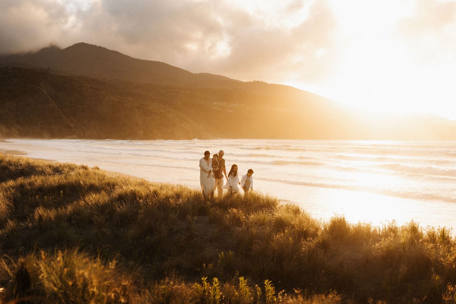 Silhouettes of people walking along a scenic beach at golden sunset with mountains in the background.