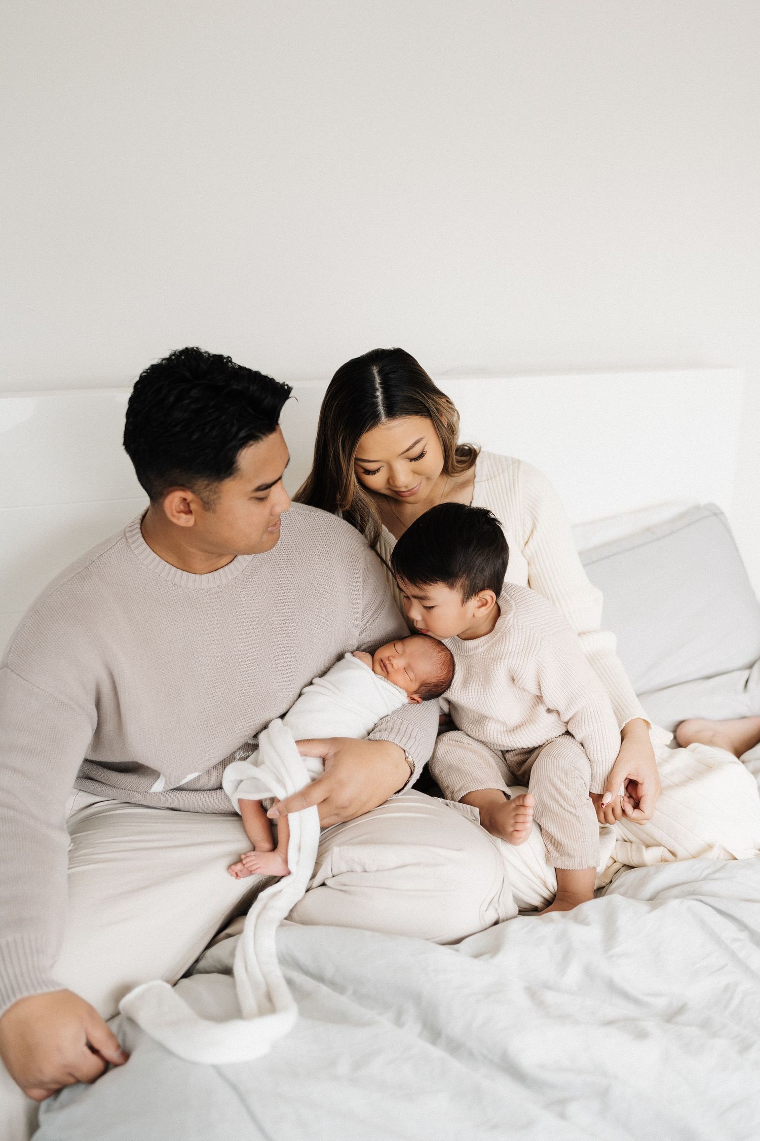 A family relaxes together on white bedding in a minimalist bedroom setting.