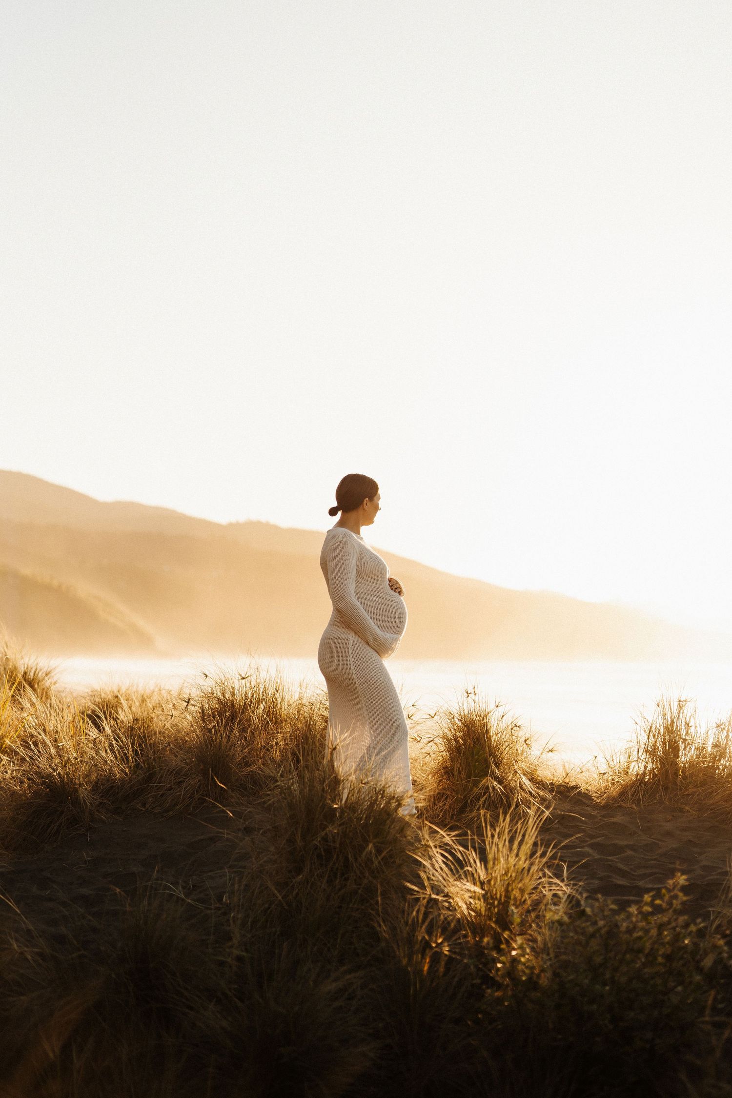A silhouette figure stands on grassy coastal dunes during a misty golden sunset against mountain silhouettes.