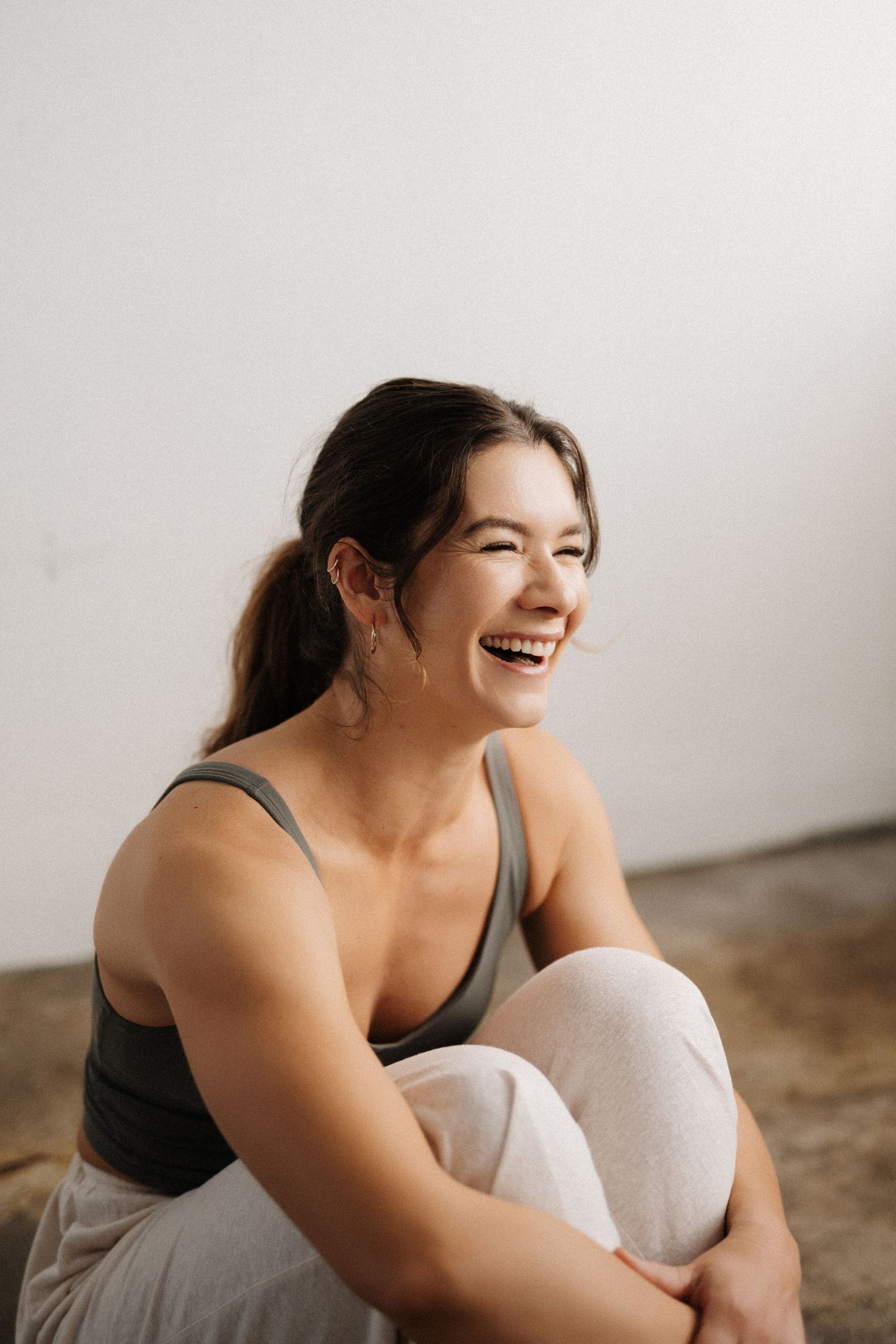 A joyful person in a gray tank top sits casually against a white wall, laughing warmly.