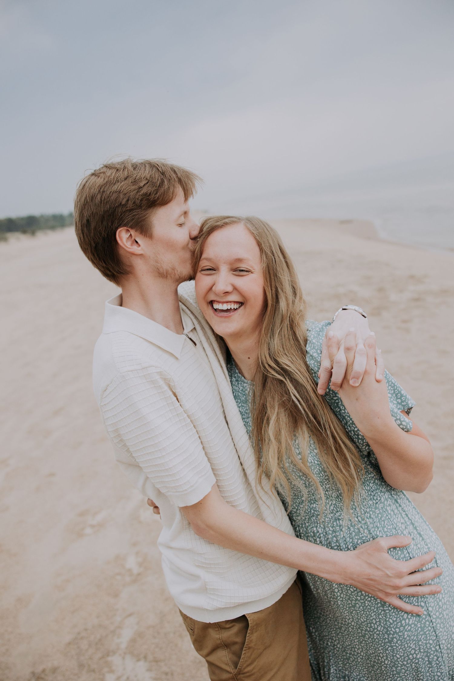 A joyful couple embraces on a sandy beach, the man kissing her cheek as she laughs, wearing a floral dress.