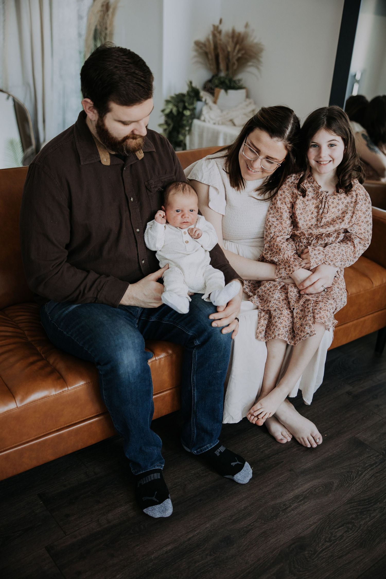 A bearded father holds a newborn baby while mother and young daughter smile beside him on a brown leather couch.