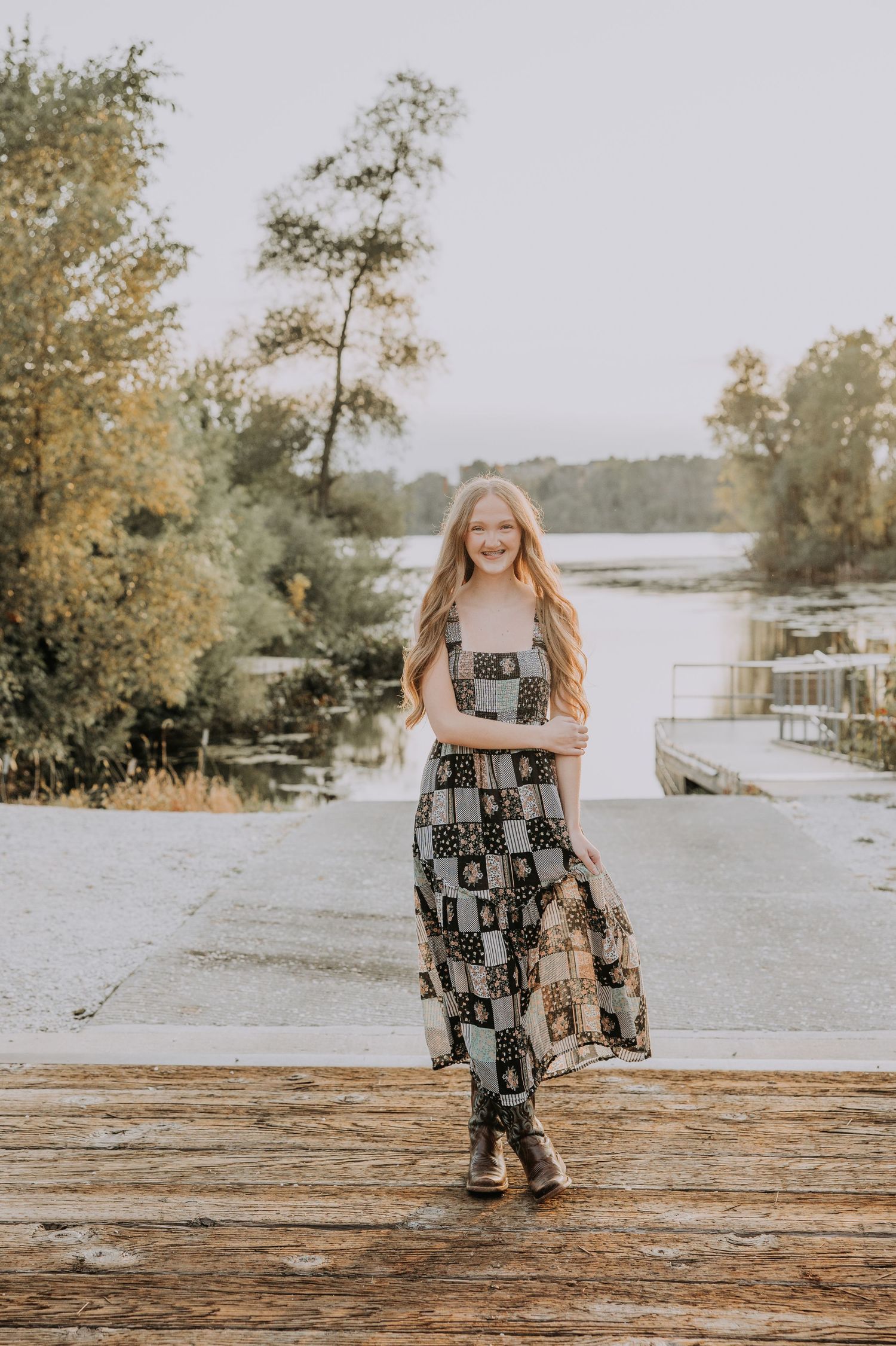 Young girl in patchwork maxi dress and boots smiling on wooden dock by lake surrounded by autumn trees.