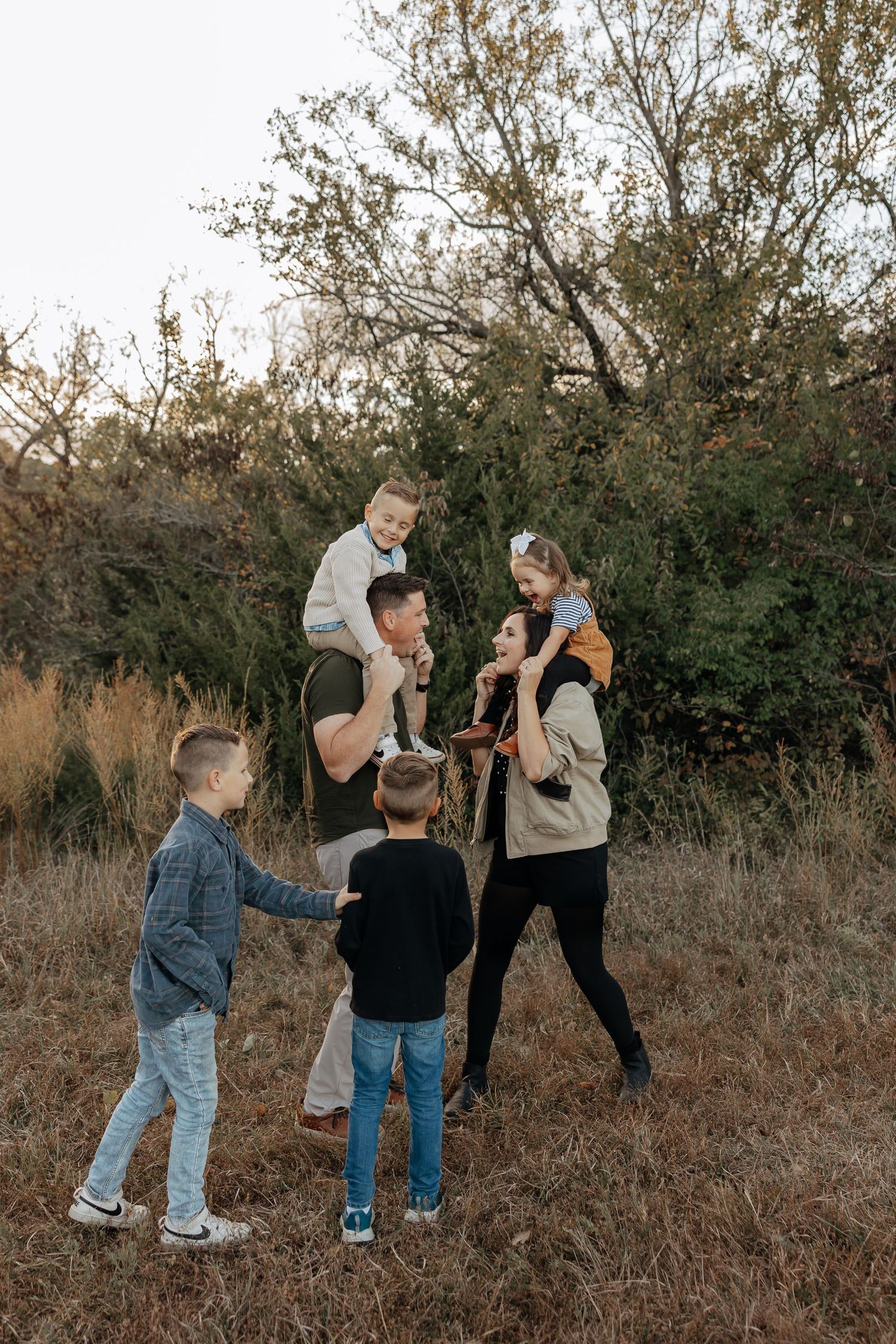 Happy family of six playing outdoors in autumn field with children on parents shoulders.