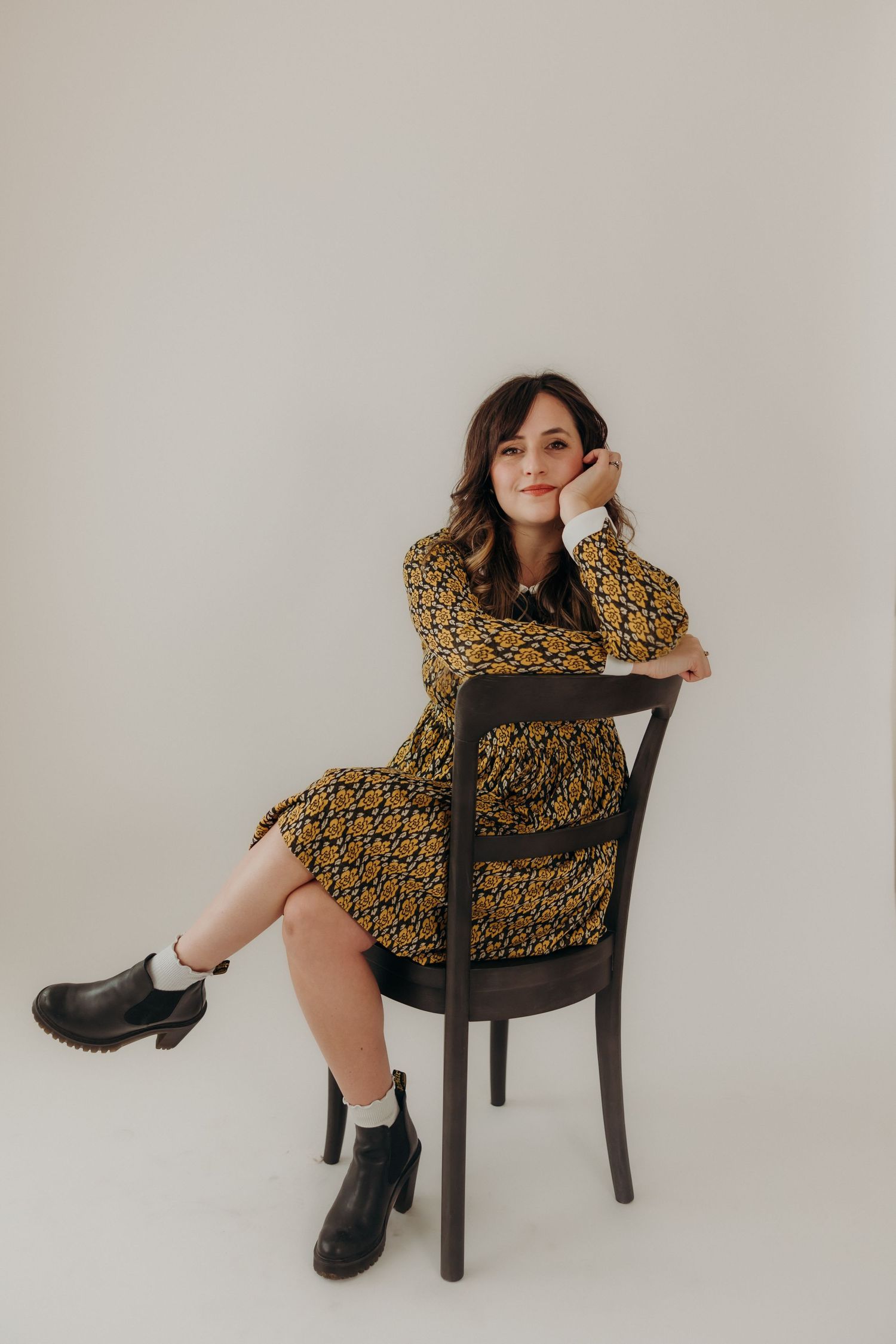 Woman in patterned yellow midi dress seated on wooden chair posing in bright white photo studio.