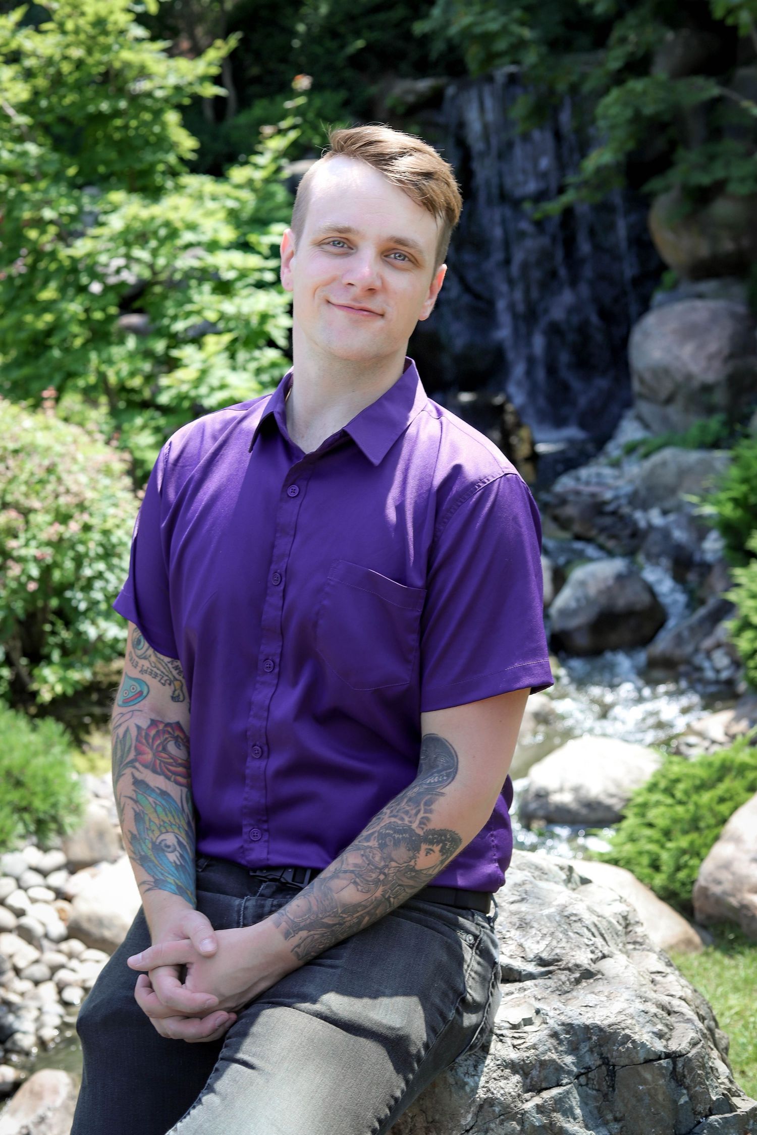 A person in a purple polo shirt with tattoos sitting on rocks near a waterfall.