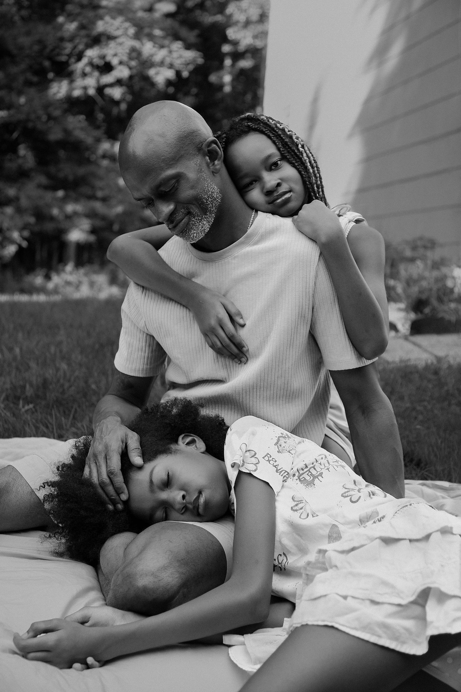 Hudson Valley family photo: dad sitting on a blanket in the backyard, one daughter hugging him from behind, another resting on his lap